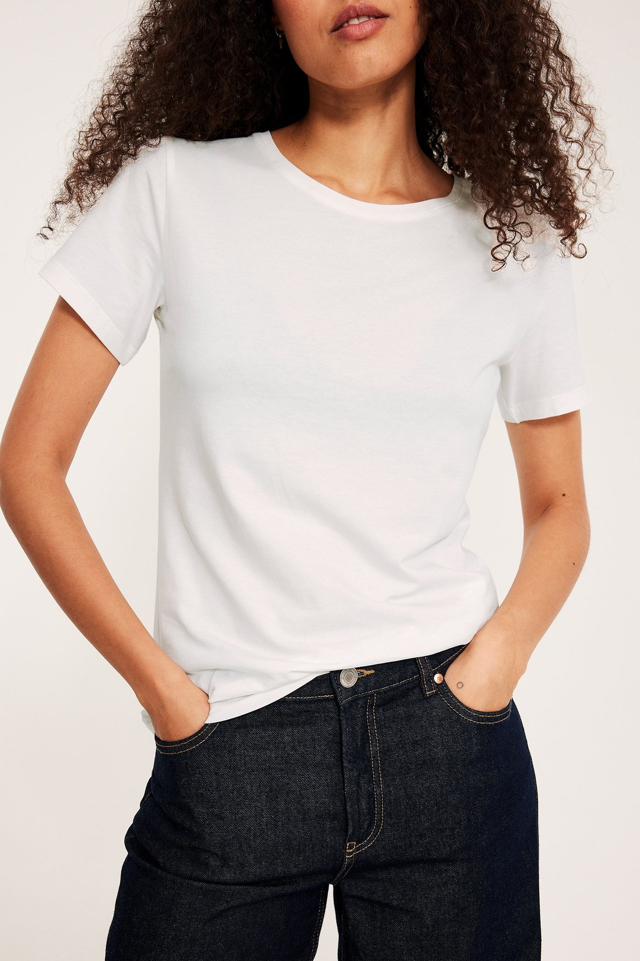 Forfra, kvinne i en offwhite kortermet basetopp med rund hals og mørkeblå jeans.