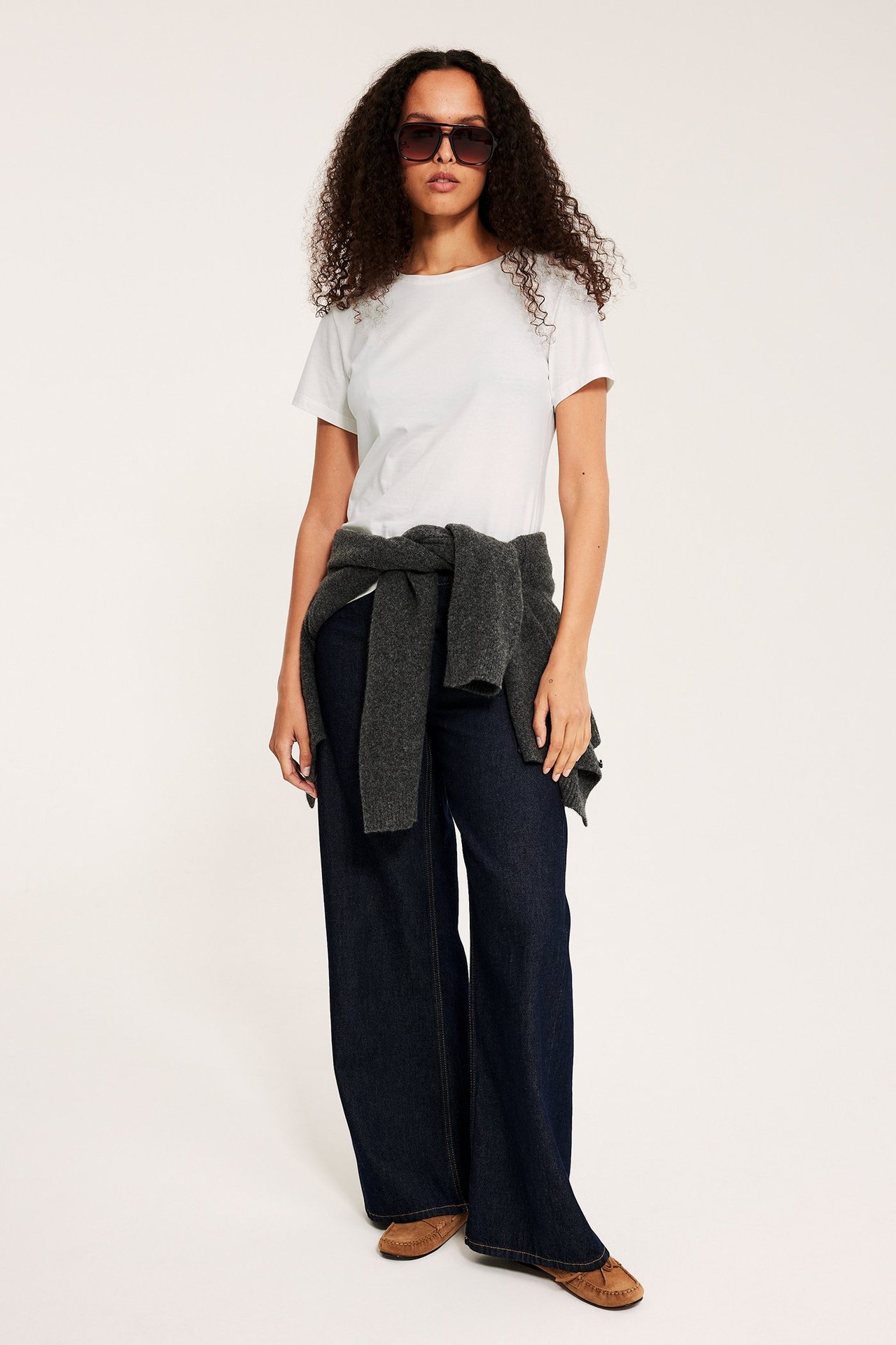 Forfra: Kvinne i offwhite kortermet topp, mørkeblå vide jeans, grå genser knyttet i midjen, solbriller.