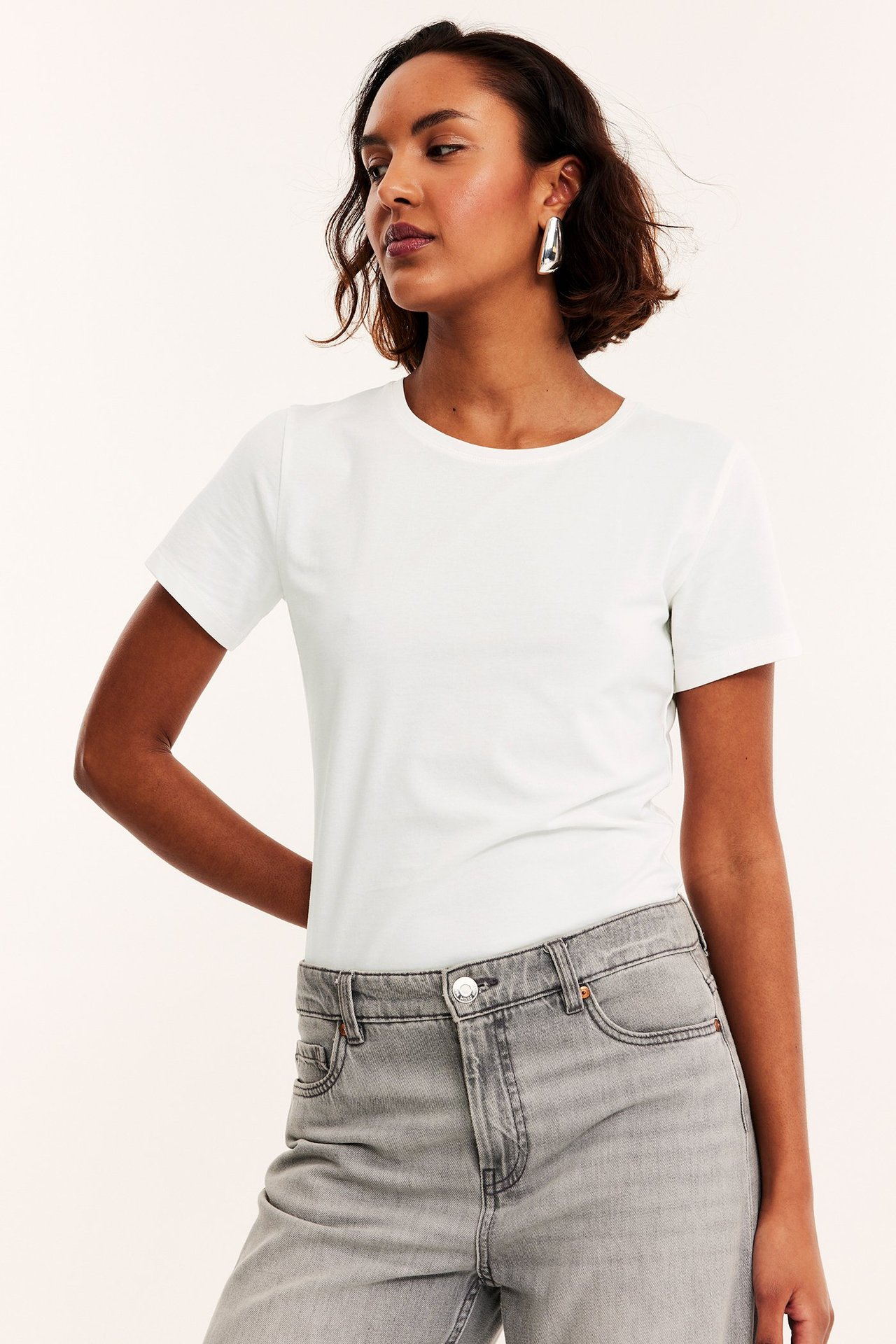 Frontbilde av kvinne i offwhite kortermet rundhalset basetopp, lysegrå jeans og sølvfargede øreringer.