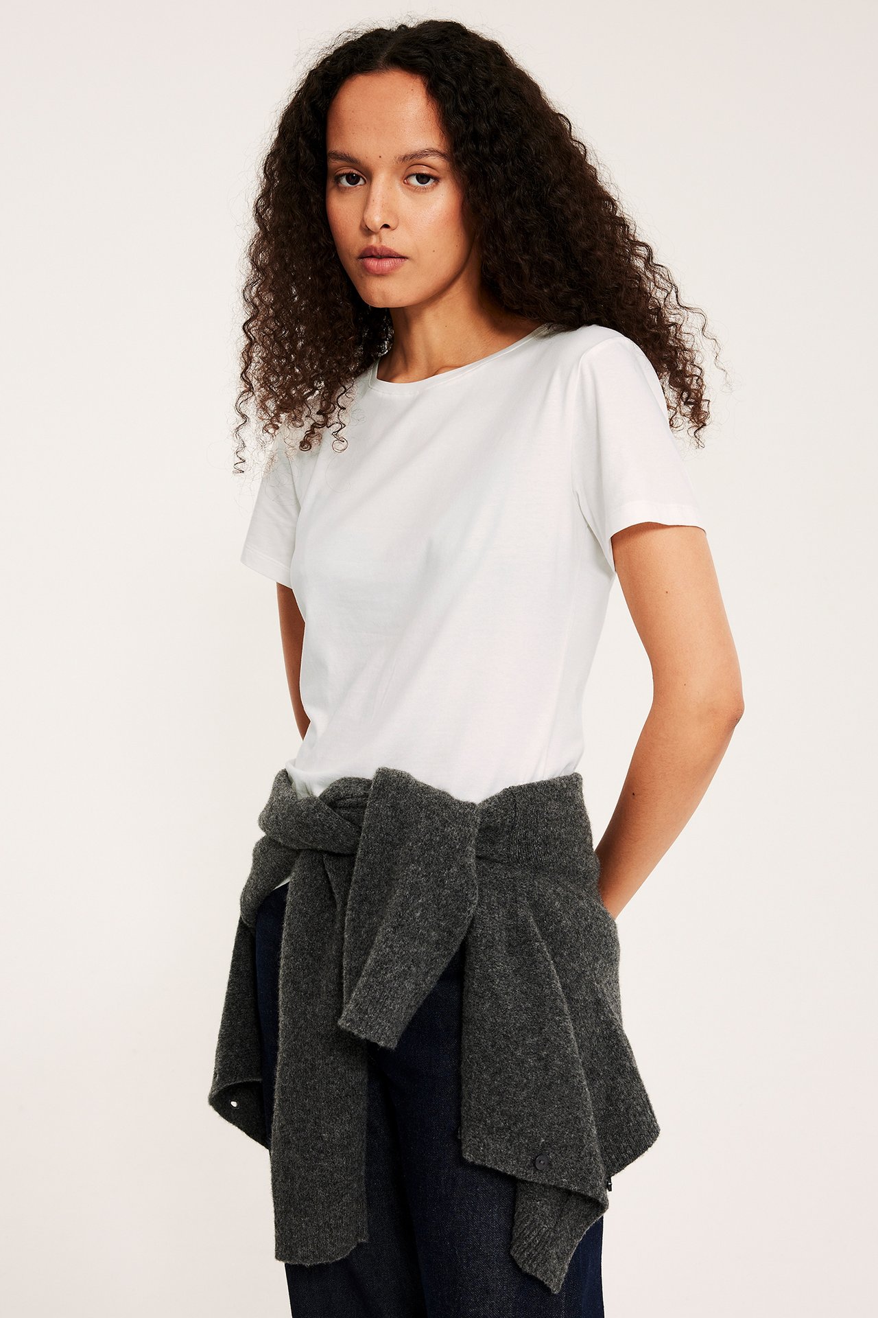 Naisen etukuva yllään luonnonvalkoinen lyhythihainen peruspaita, tummanharmaa neule vyötäröllä.