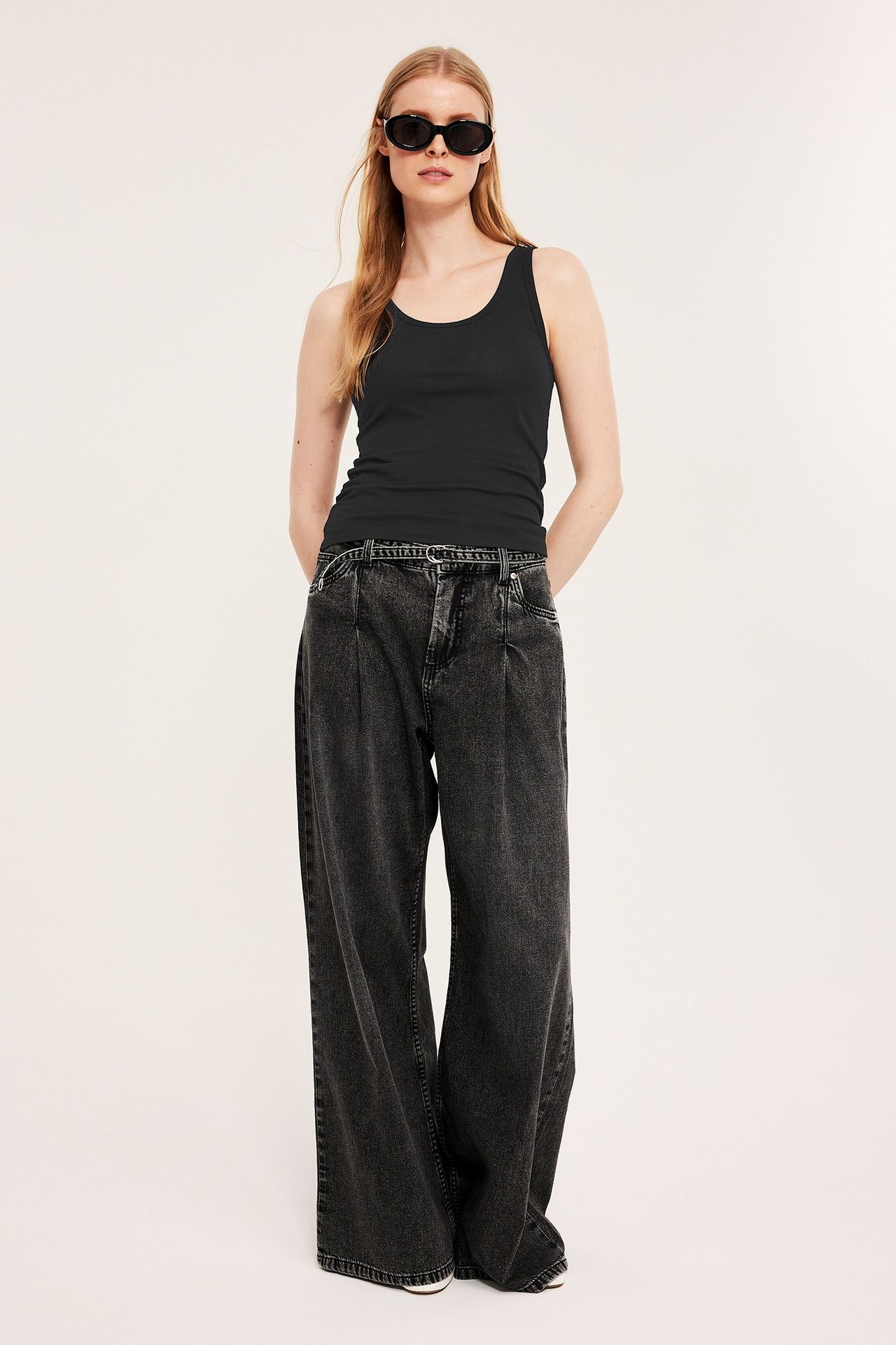 Frontbilde av en kvinne i svart stretch-topp, mørkegrå vide jeans og svarte ovale solbriller.