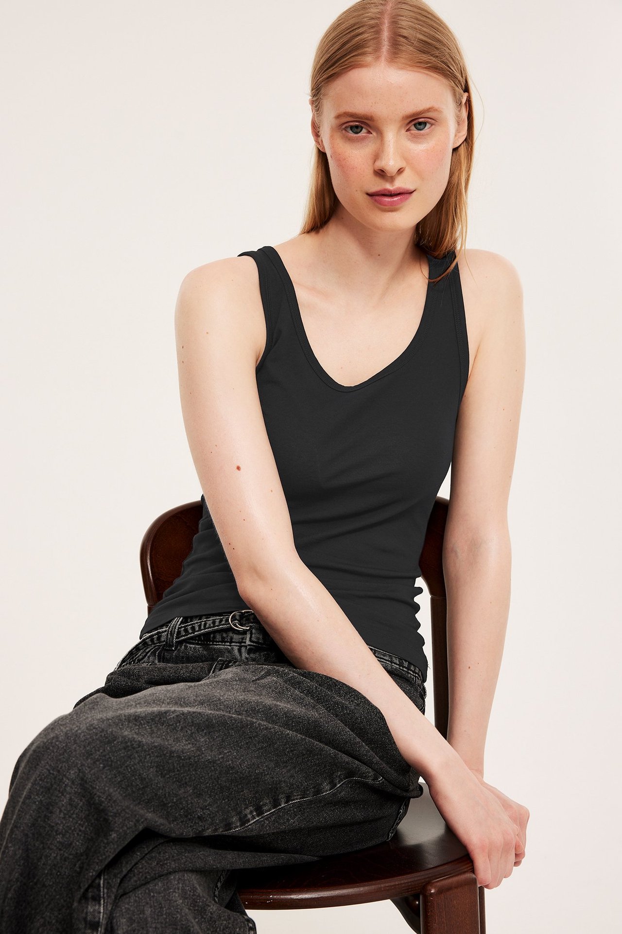 Frontbilde av en kvinne i svart tettsittende basistopp og mørkegrå jeans, sittende på en stol.