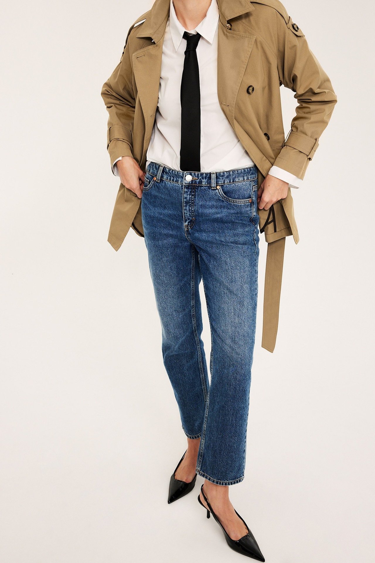 Nainen edestäpäin: siniset mid-waist suorat farkut, valkoinen paita, musta solmio ja beige trenssitakki.