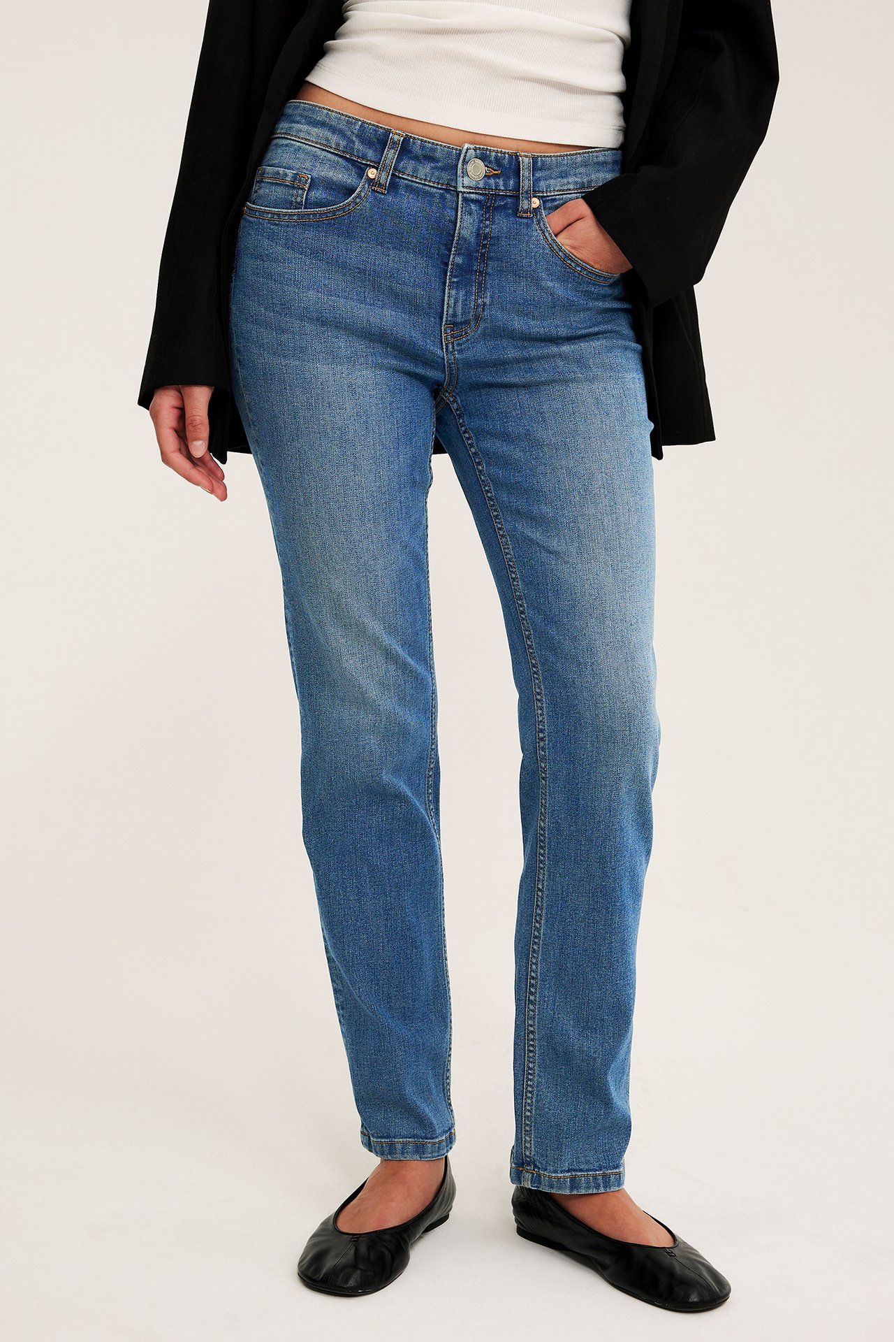 Kvinna i mellanblå raka jeans, vit topp, svart kavaj och svarta ballerinaskor, framifrån.