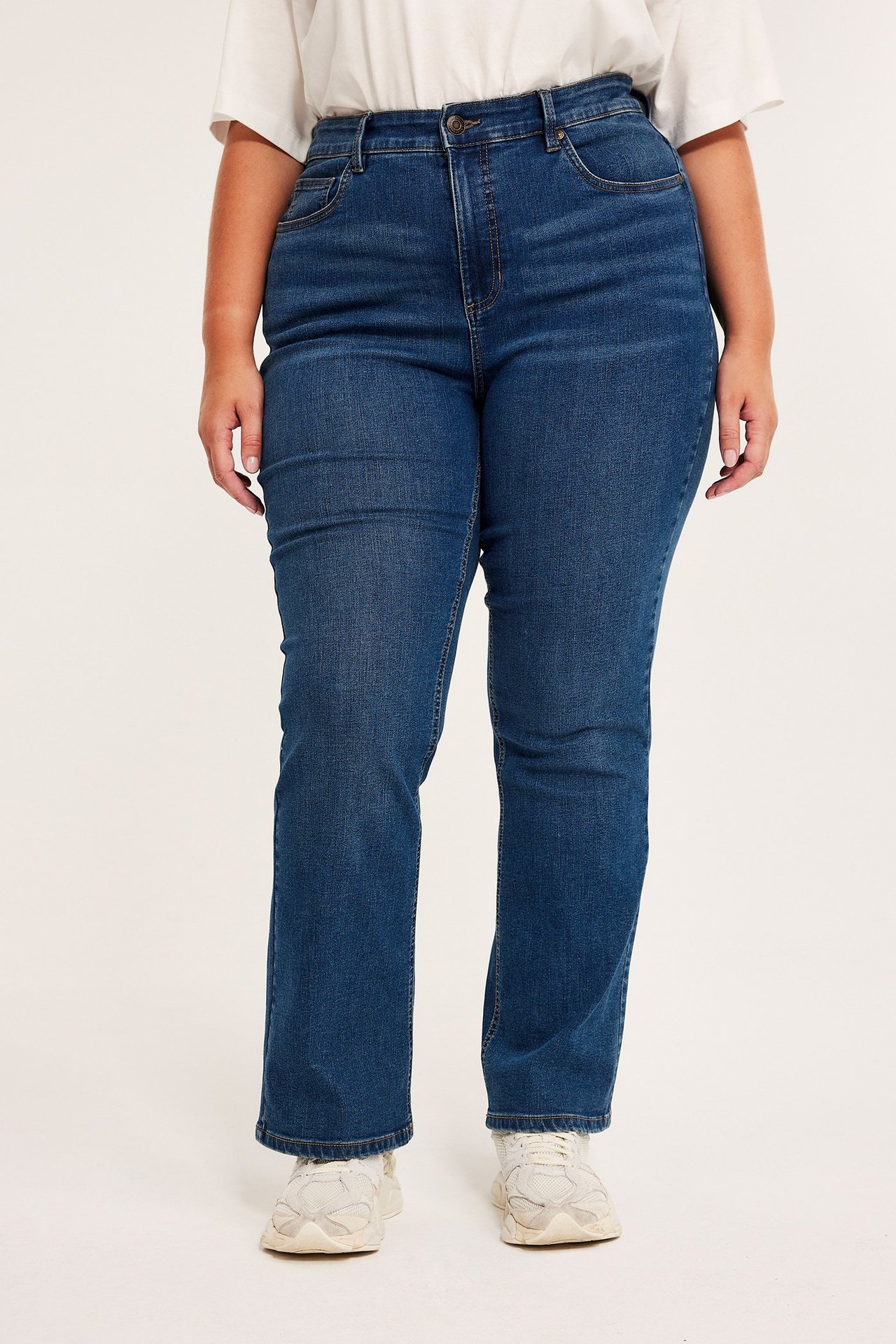 Edestäpäin: nainen yllään korkeavyötäröiset siniset bootcut-farkut, valkoinen t-paita ja valkoiset lenkkarit.
