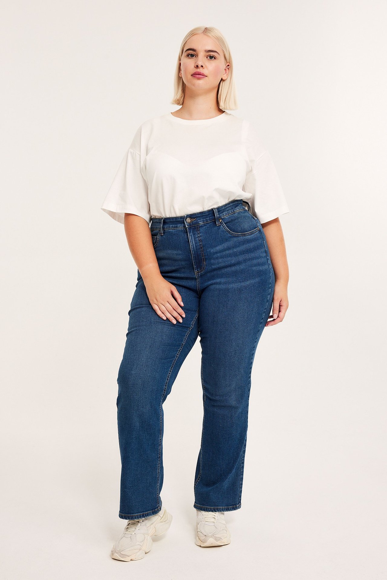 Edestä kuvattu nainen yllään korkeavyötäröiset siniset denim bootcut-farkut, valkoinen oversized t-paita ja beiget lenkkarit.
