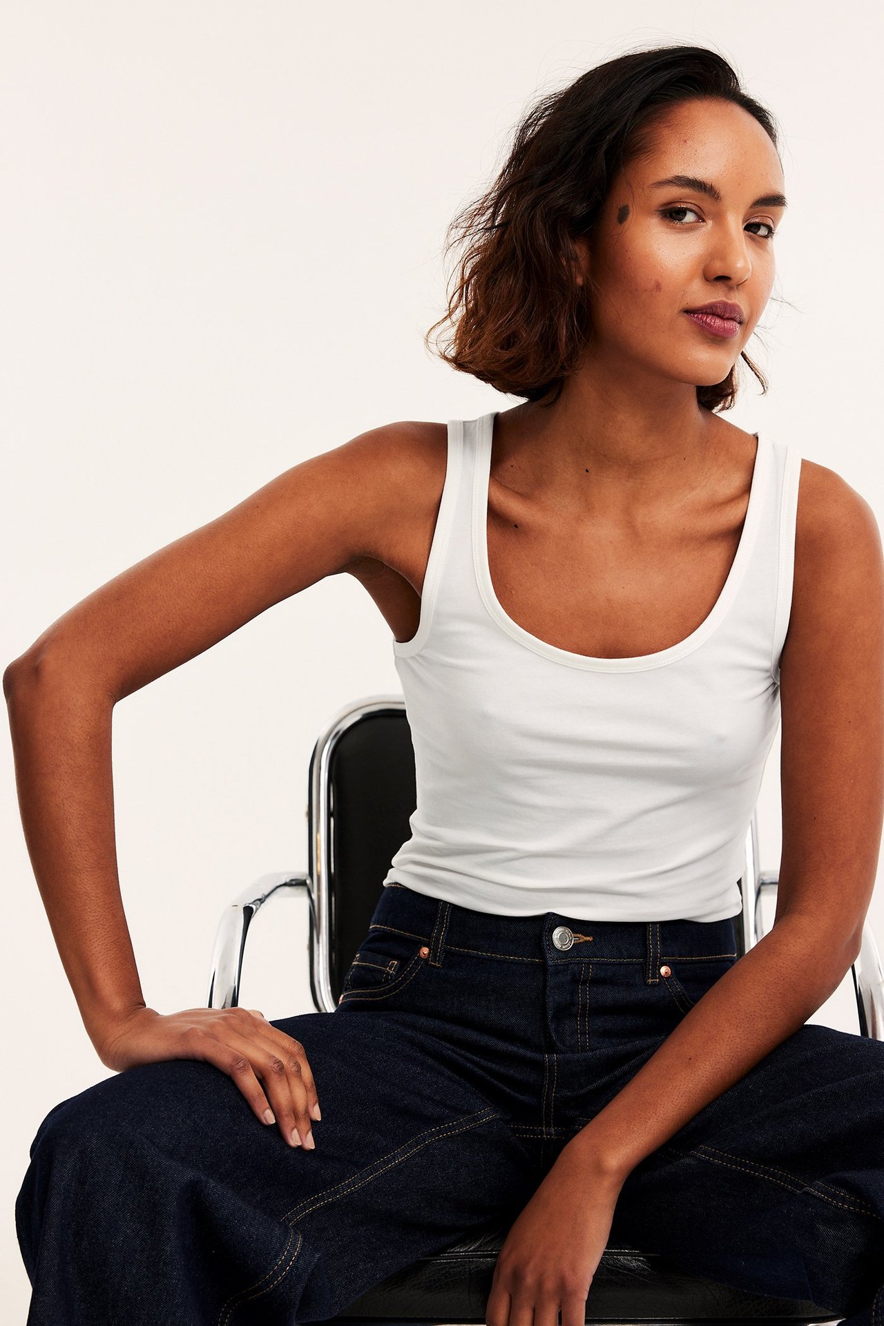 Frontvisning av en kvinne i en offwhite basic stretch singlet og mørkeblå vide jeans.