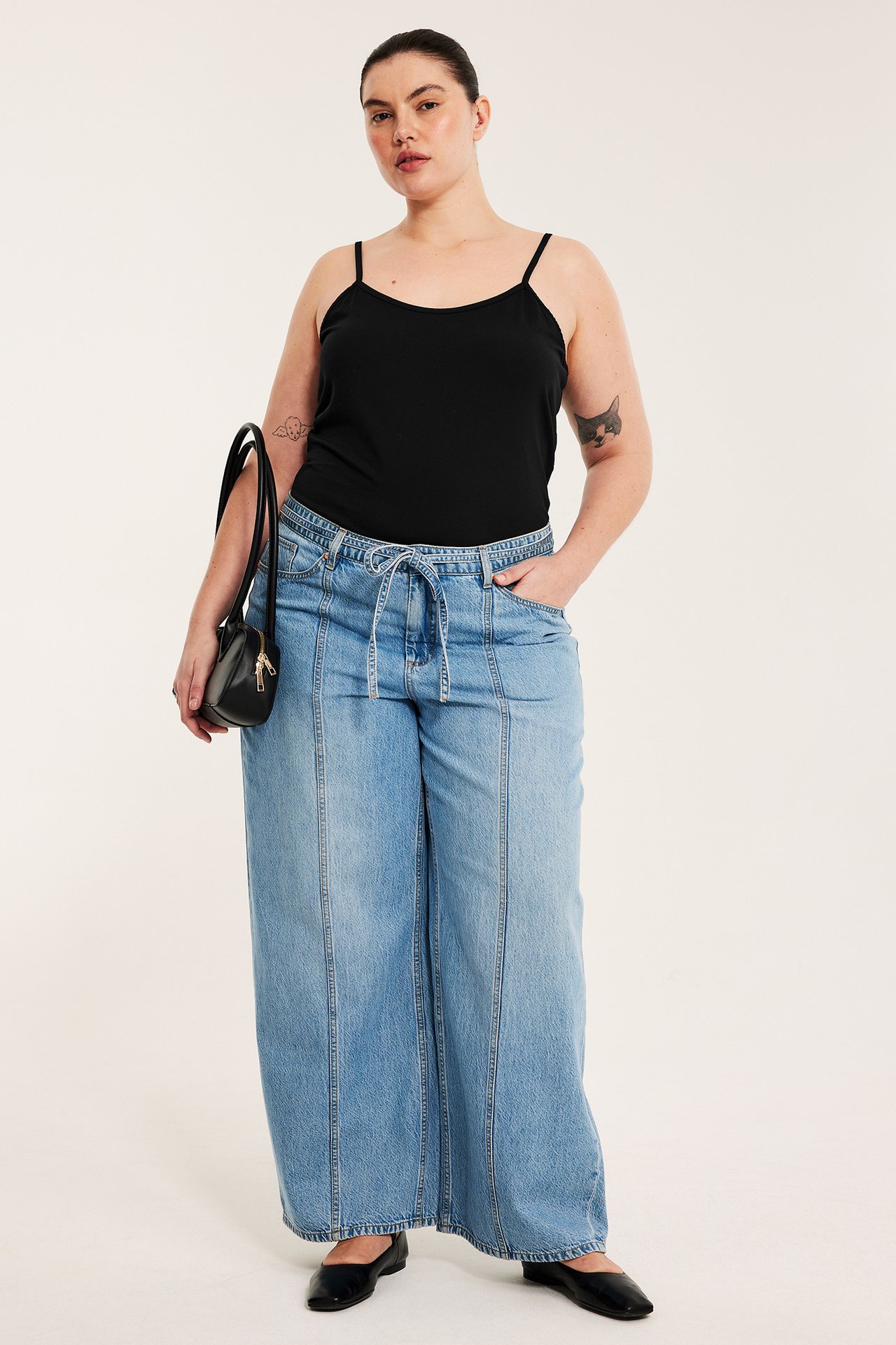 Forfra: kvinne i svart jerseylinne, lyseblå vide jeans, svarte ballerinasko, holder en svart veske.