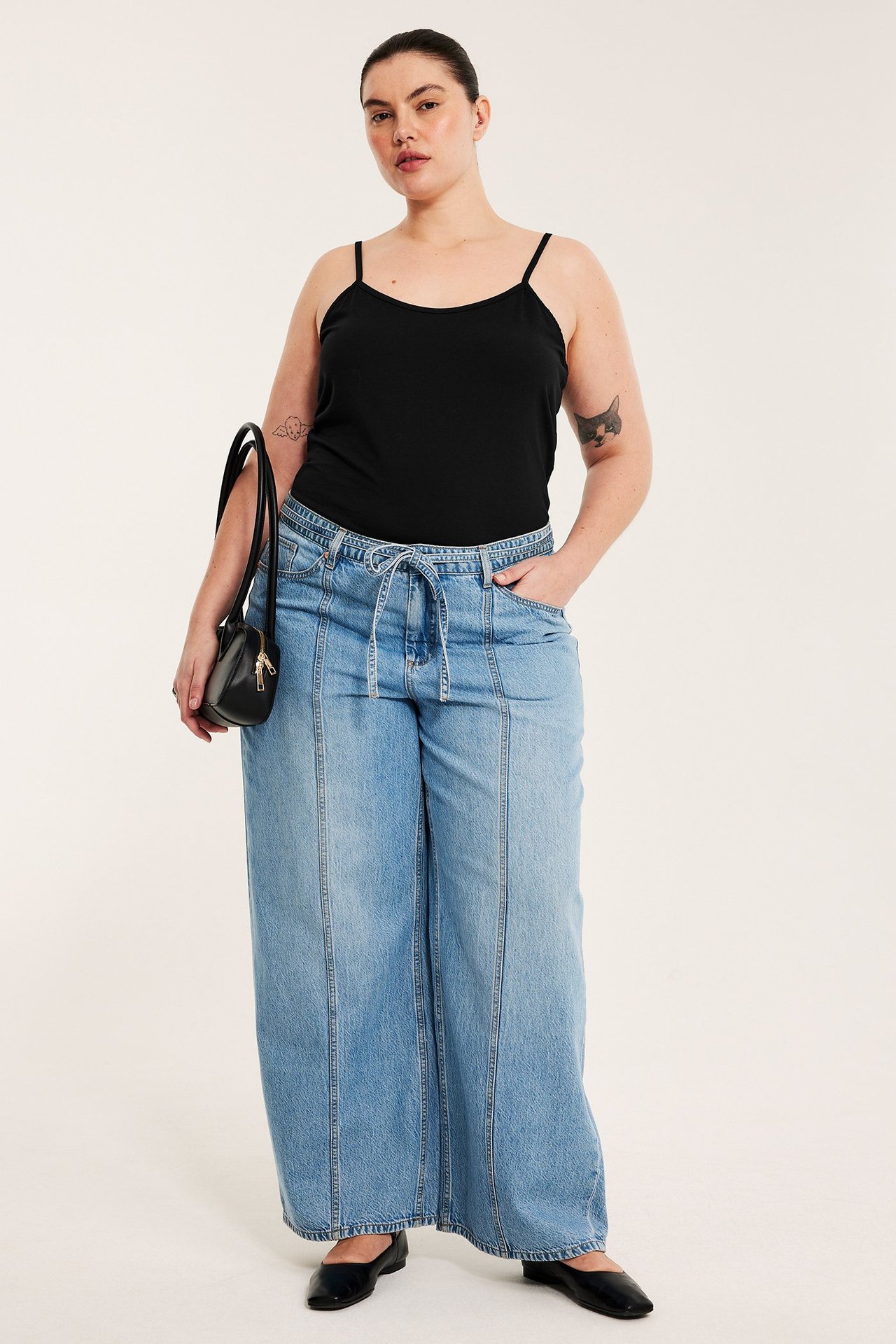 Forfra: kvinne i svart jerseylinne, lyseblå vide jeans, svarte ballerinasko, holder en svart veske.