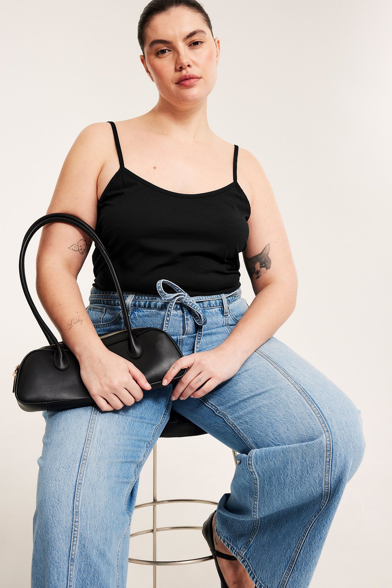 Frontbilde av sittende kvinne i svart jerseylinne og lyseblå vide jeans, holdende en svart veske.