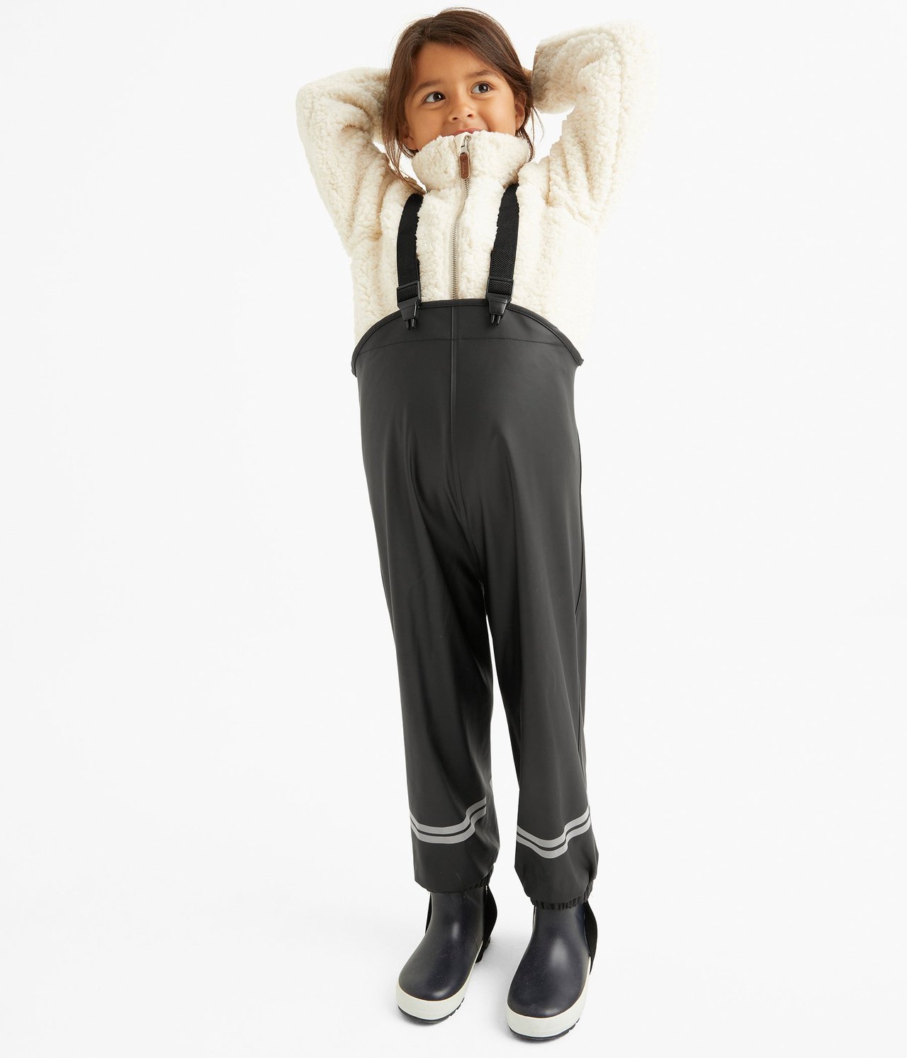Framsida av ett barn i mörkgrå regnbyxor med hängslen, reflexränder, gräddvit fleecejacka och mörkblå stövlar.