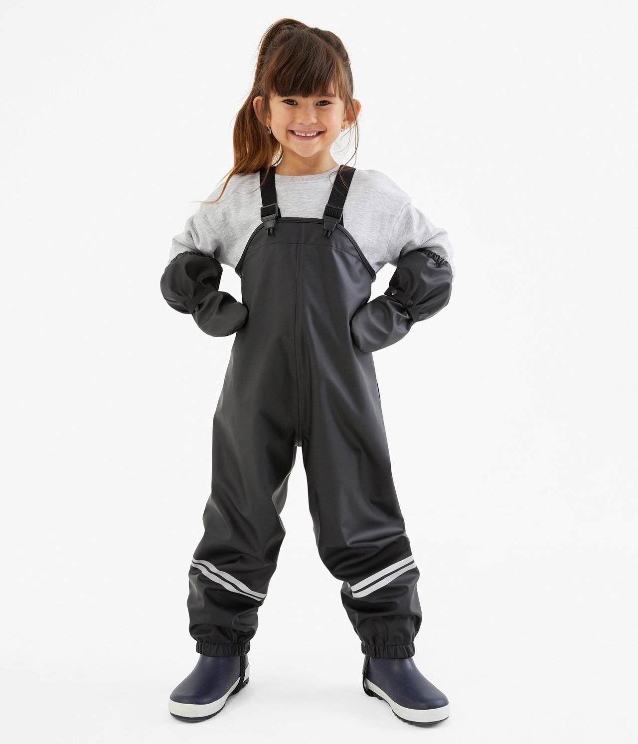 Barn i svarta regnbyxor med hängslen, reflexer, grå tröja, svarta vantar och blå stövlar.