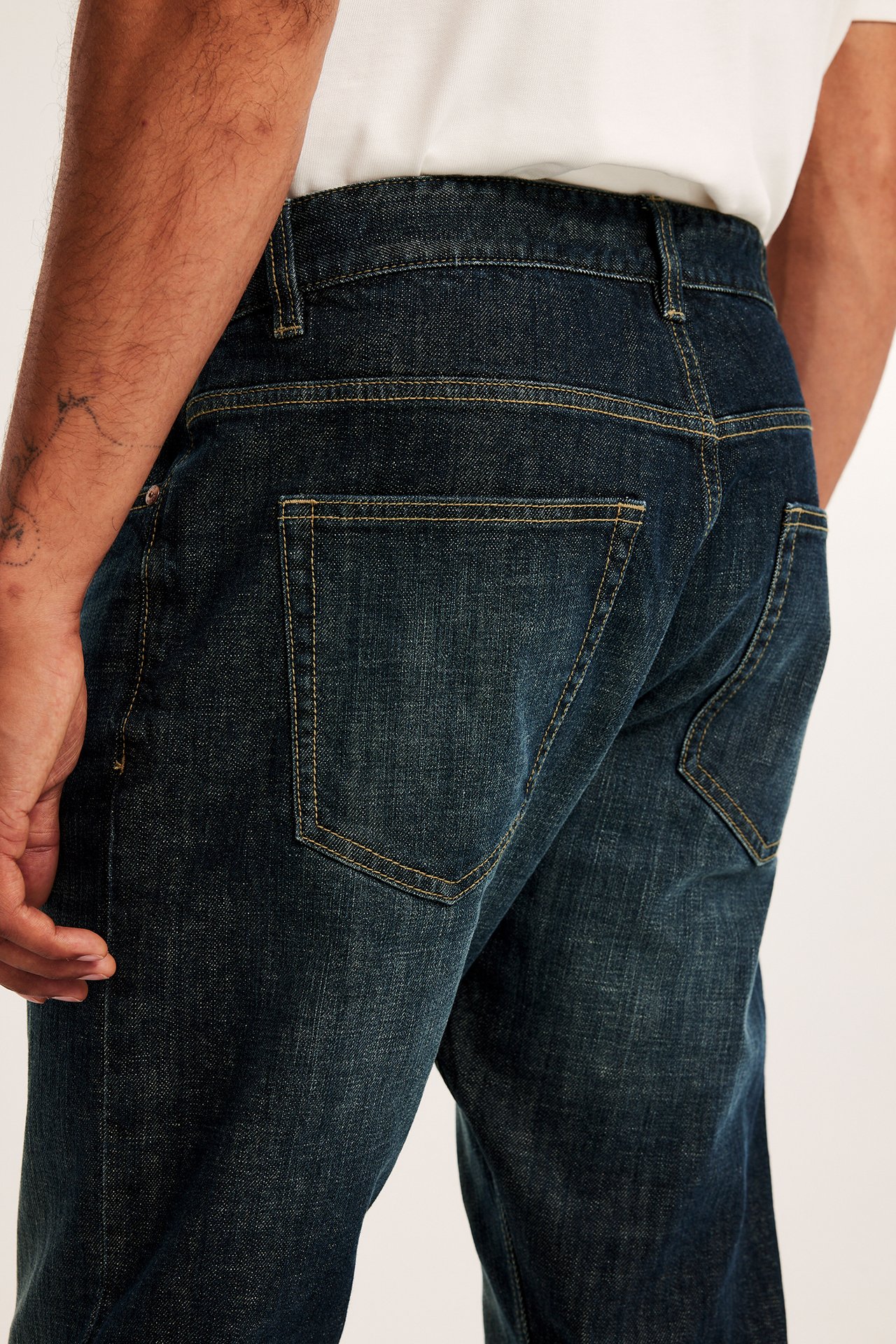 Bakifrån, man i mörkblå regular fit denimjeans med gula sömmar och en vit t-shirt.
