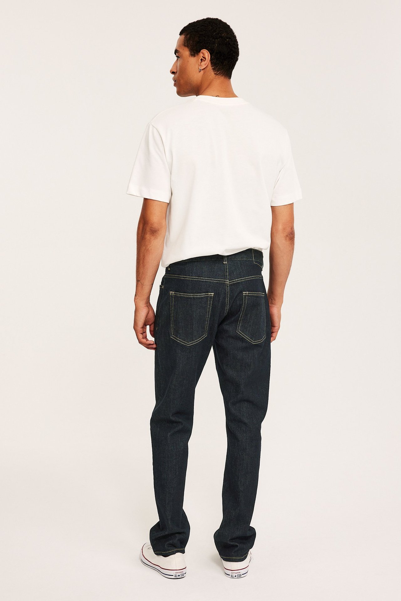 Bakifrån, en man i mörk denim regular fit jeans med gula sömmar, en vit t-shirt och vita sneakers.