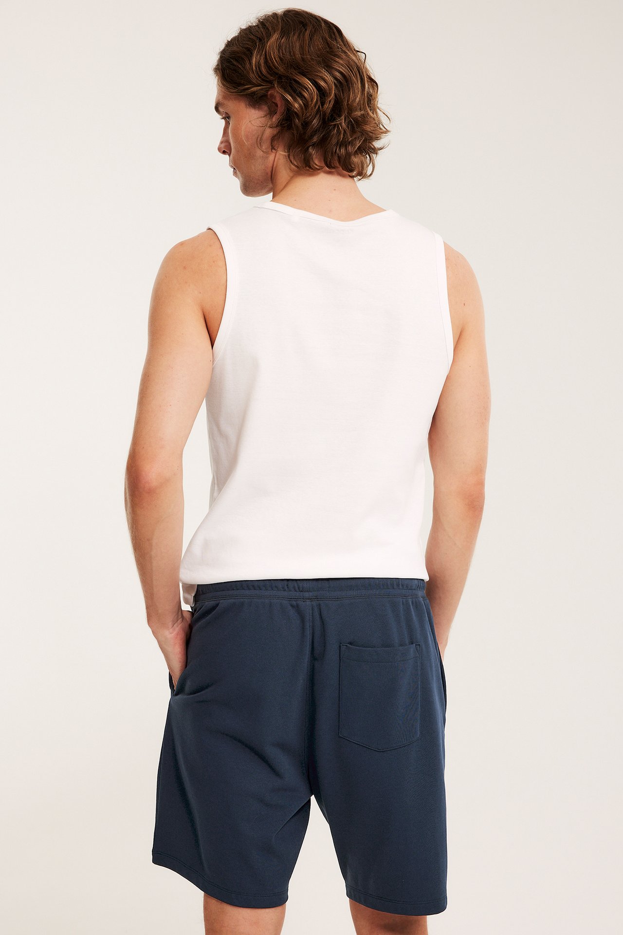 Mies takaa: tummansiniset college-shortsit joustavalla vyötäröllä, sivutaskuilla ja valkoinen hihaton paita.