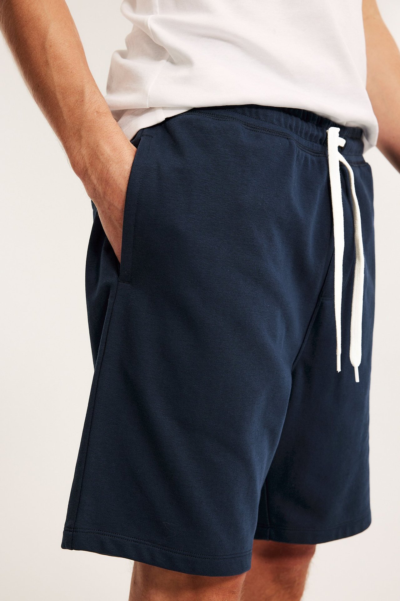 Lähikuva miehestä yllään tummansiniset college-shortsit, joissa valkoinen kiristysnyöri, sivutaskut ja valkoinen t-paita.