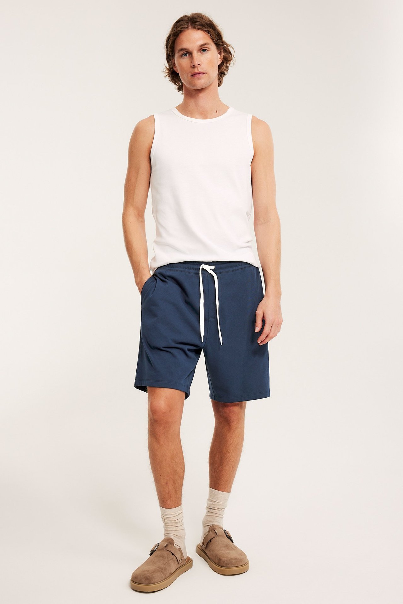 Man i mörkblå mjukisshorts med vit dragsko, vitt linne, beiga strumpor och bruna tofflor.