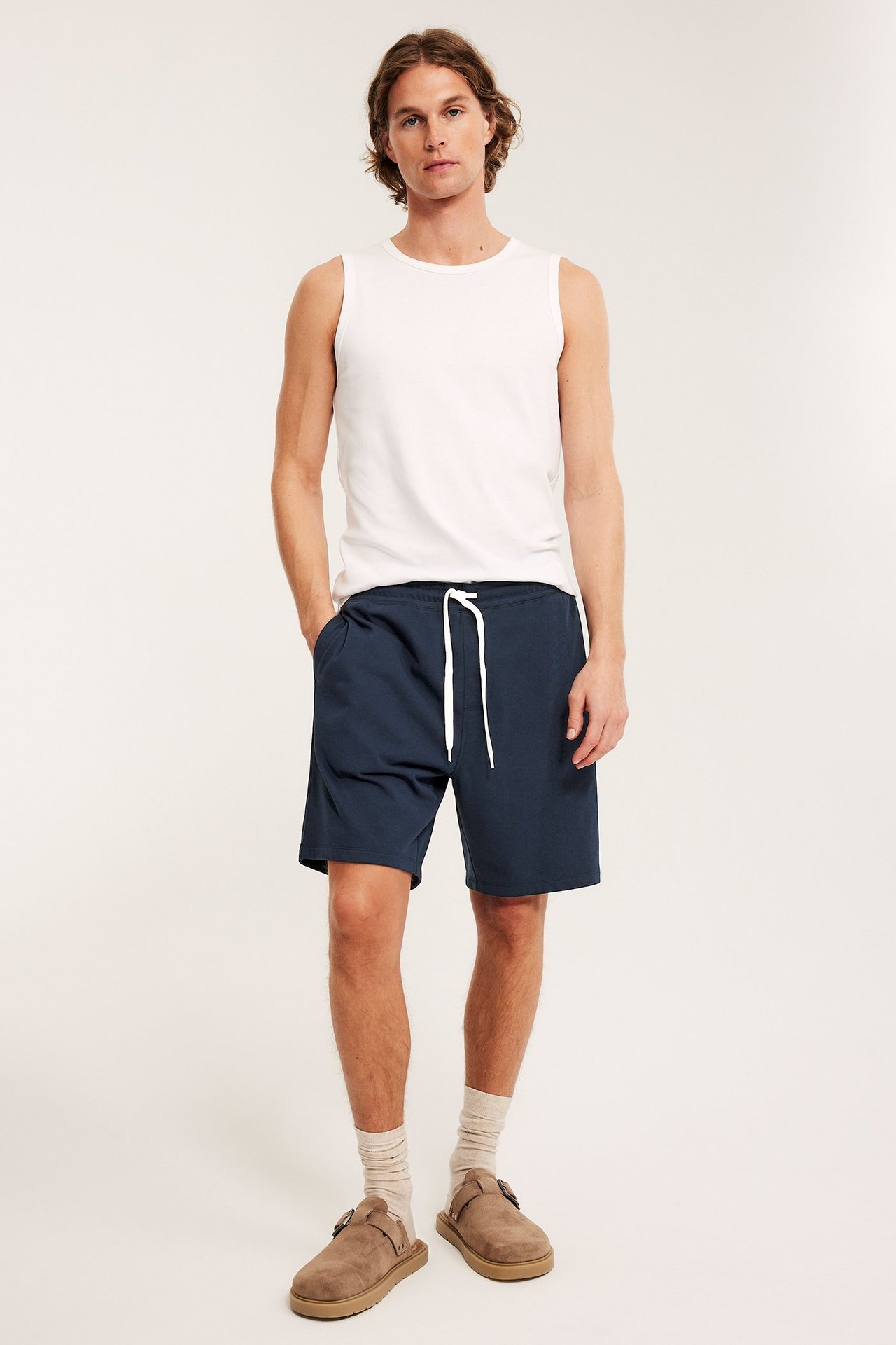 Mężczyzna w granatowych szortach dresowych z białym sznurkiem, białej koszulce, beżowych skarpetkach i brązowych klapkach.