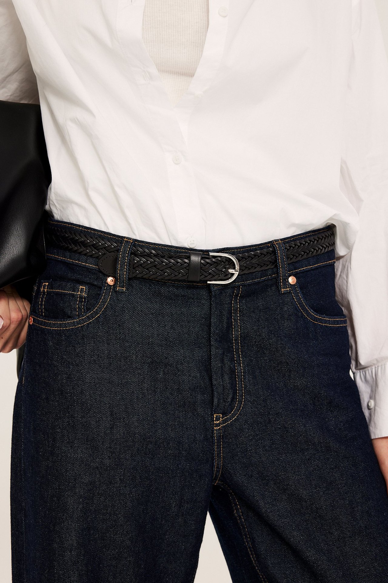 Nainen mustassa punotussa nahkavyössä hopeasoljella, tummissa farkuissa ja valkoisessa paidassa.