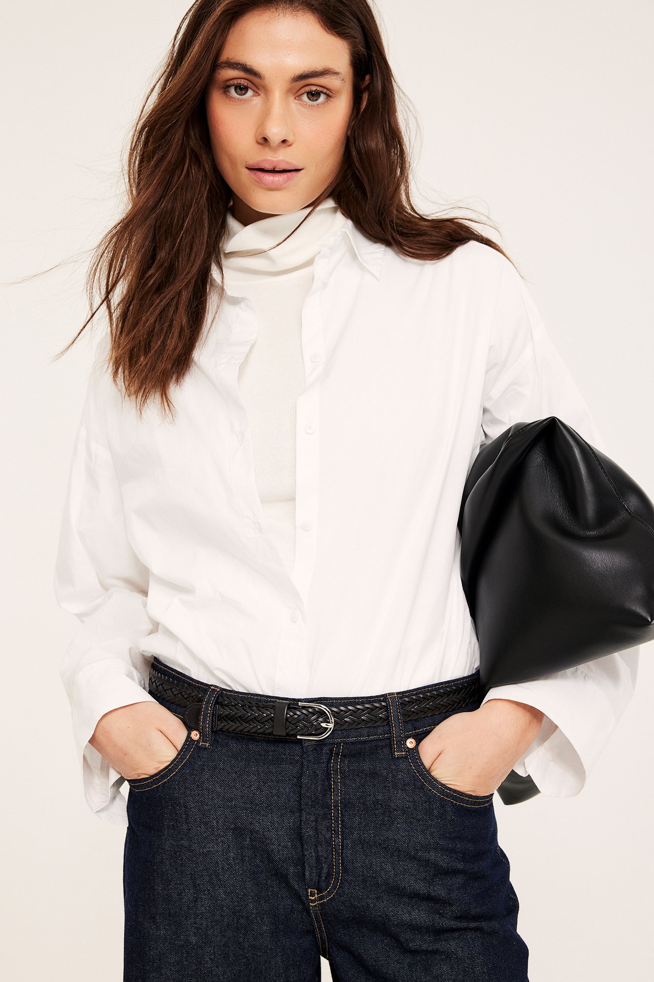 Nainen mustassa punotussa nahkavyössä, valkoisessa paidassa, tummansinisissä farkuissa, pitäen mustaa laukkua.