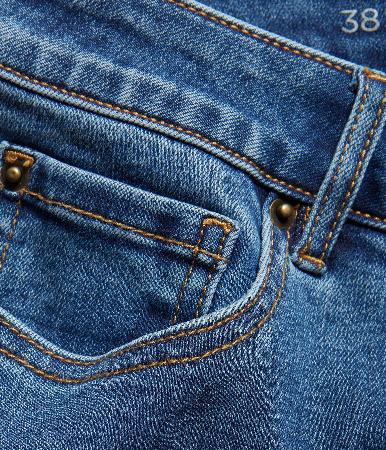 Zbliżenie na niebieskie proste dżinsy, pokazujące przednią kieszeń z pomarańczowymi szwami i mosiężnymi nitami.
