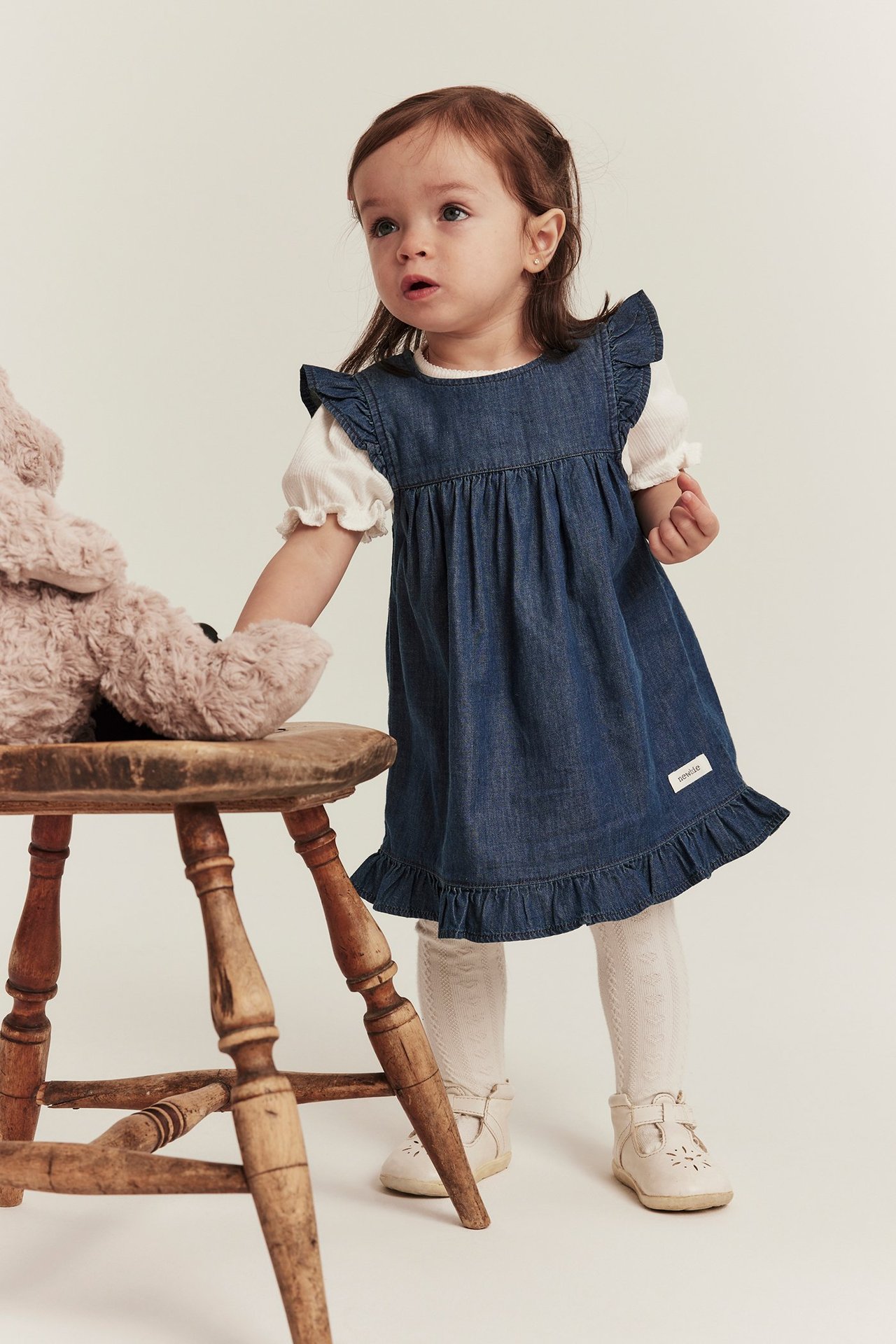 Framsida: Baby bär en jeansklänning med volang, vit ribbad topp, mönstrade strumpbyxor och beiga skor.