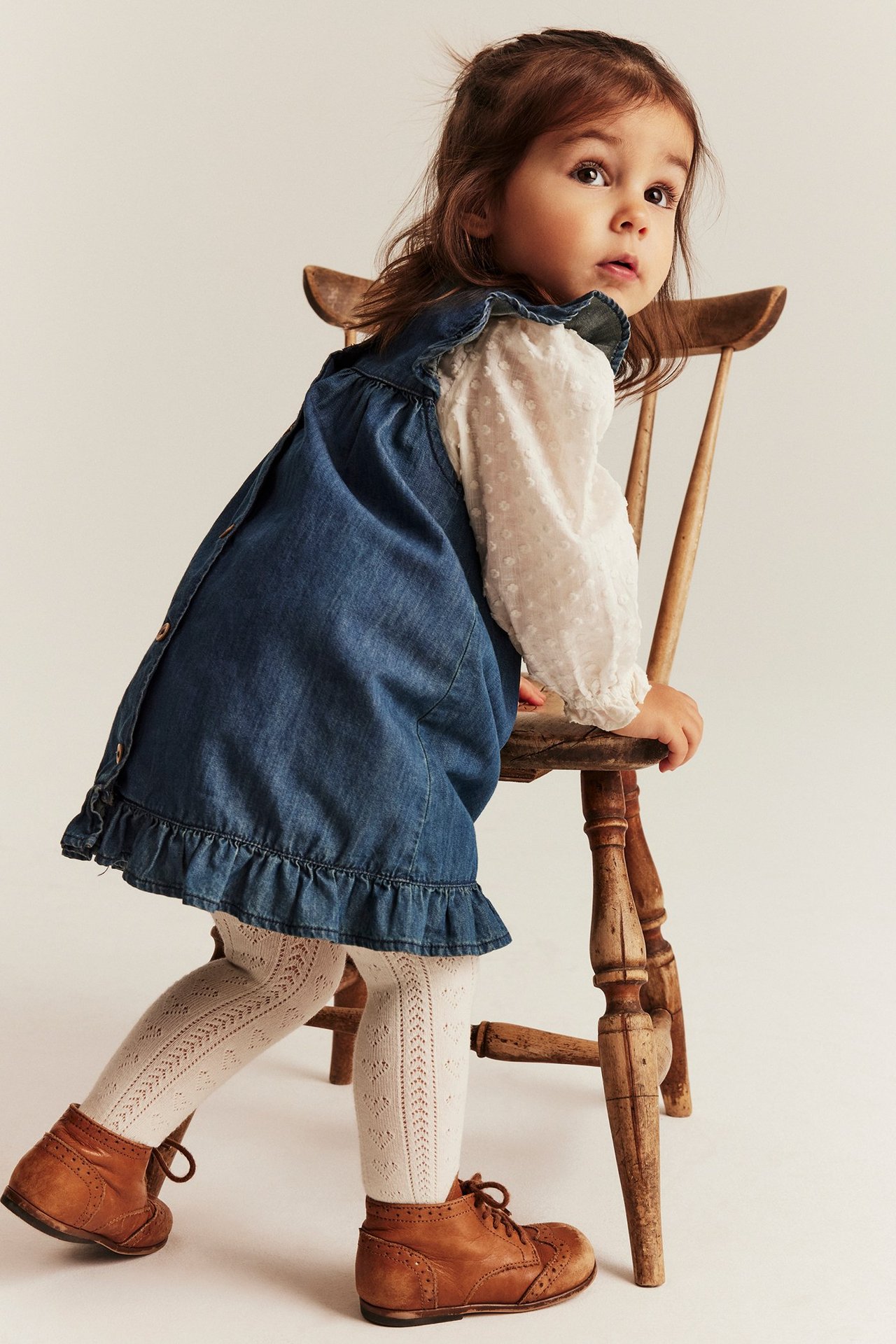 Sidebilde av et barn i blå denim-volangskjole, hvit prikket bluse, lys beige strømpebukse og brune støvler.