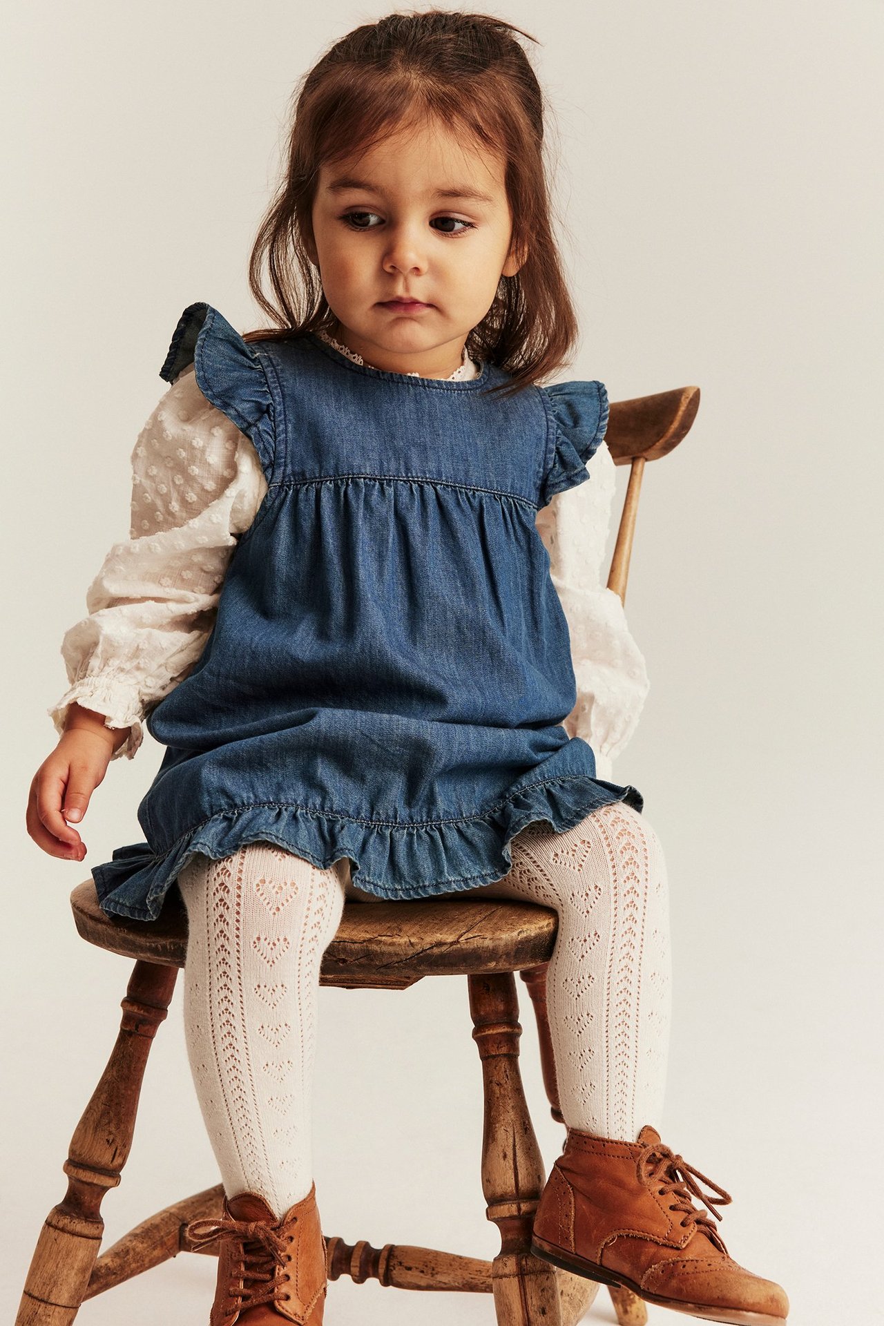 Frontbilde av baby i denimkjole med rysjer, hvit teksturert skjorte, hvite hjertestrømpebukser og brune støvler.