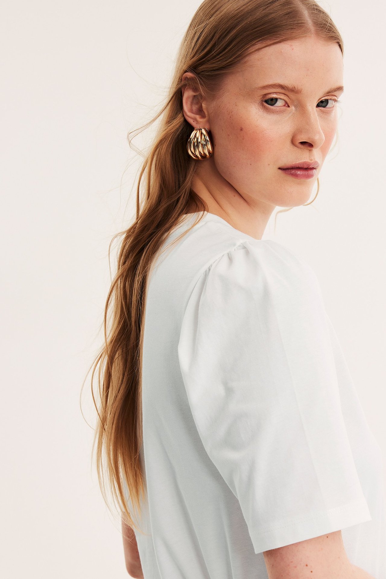 Nærbilde, sidevisning av en kvinne i offwhite bomullstopp med puffermer og gullfargede ringøreringer.