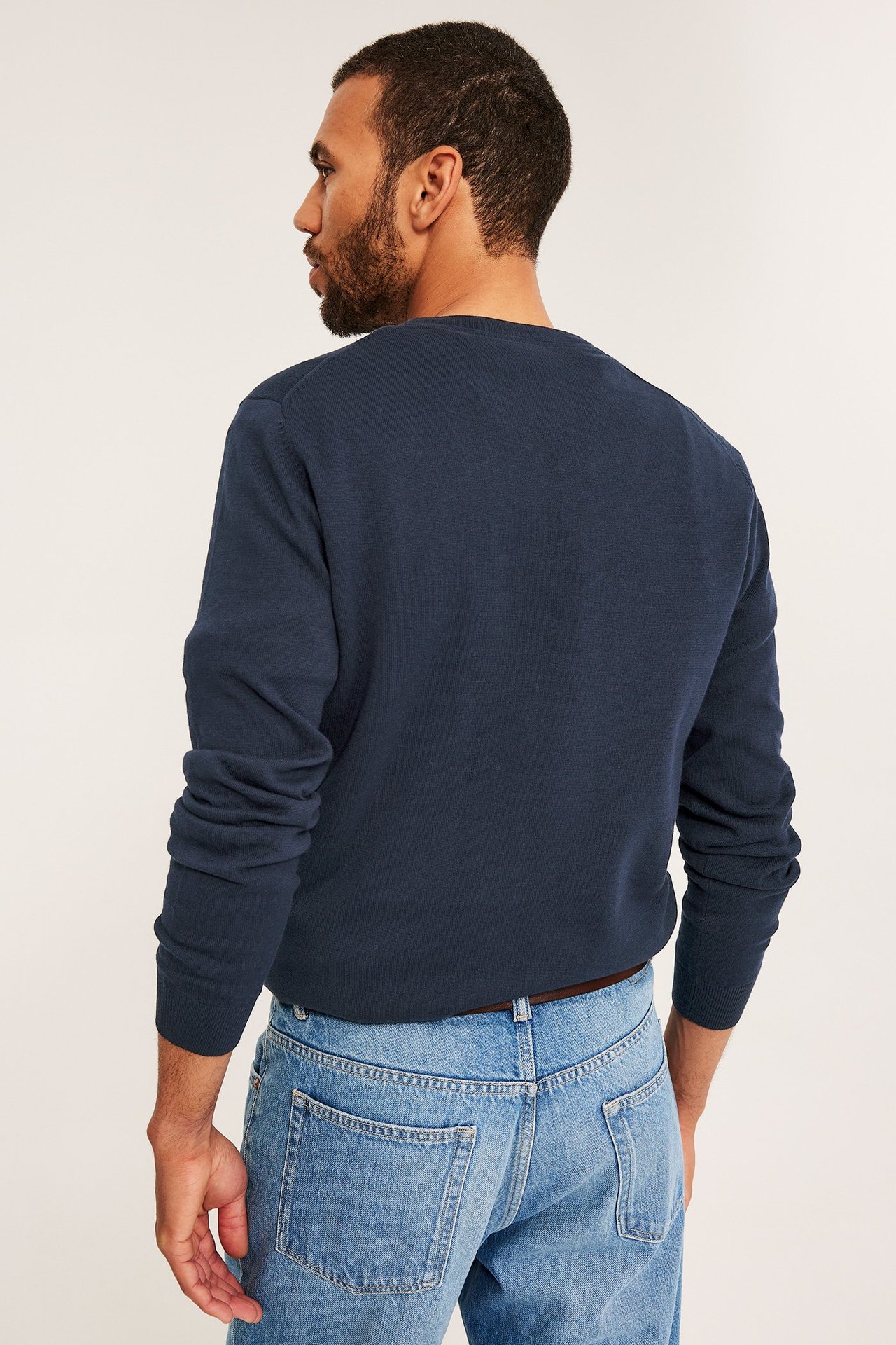 Bakifrån, en man i mörkblå finstickad bomullströja, ljusblå jeans och ett brunt bälte.