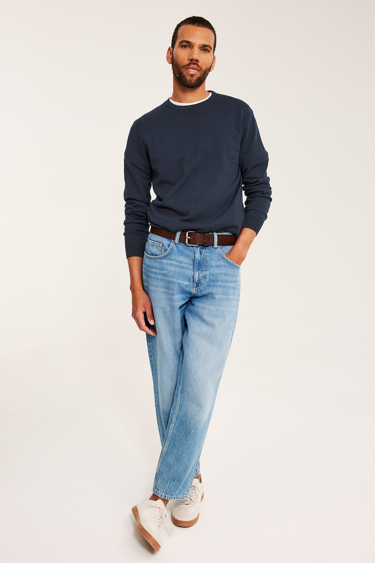 Man i mörkblå finstickad bomullströja, ljusblåa jeans, brunt bälte och vita sneakers.