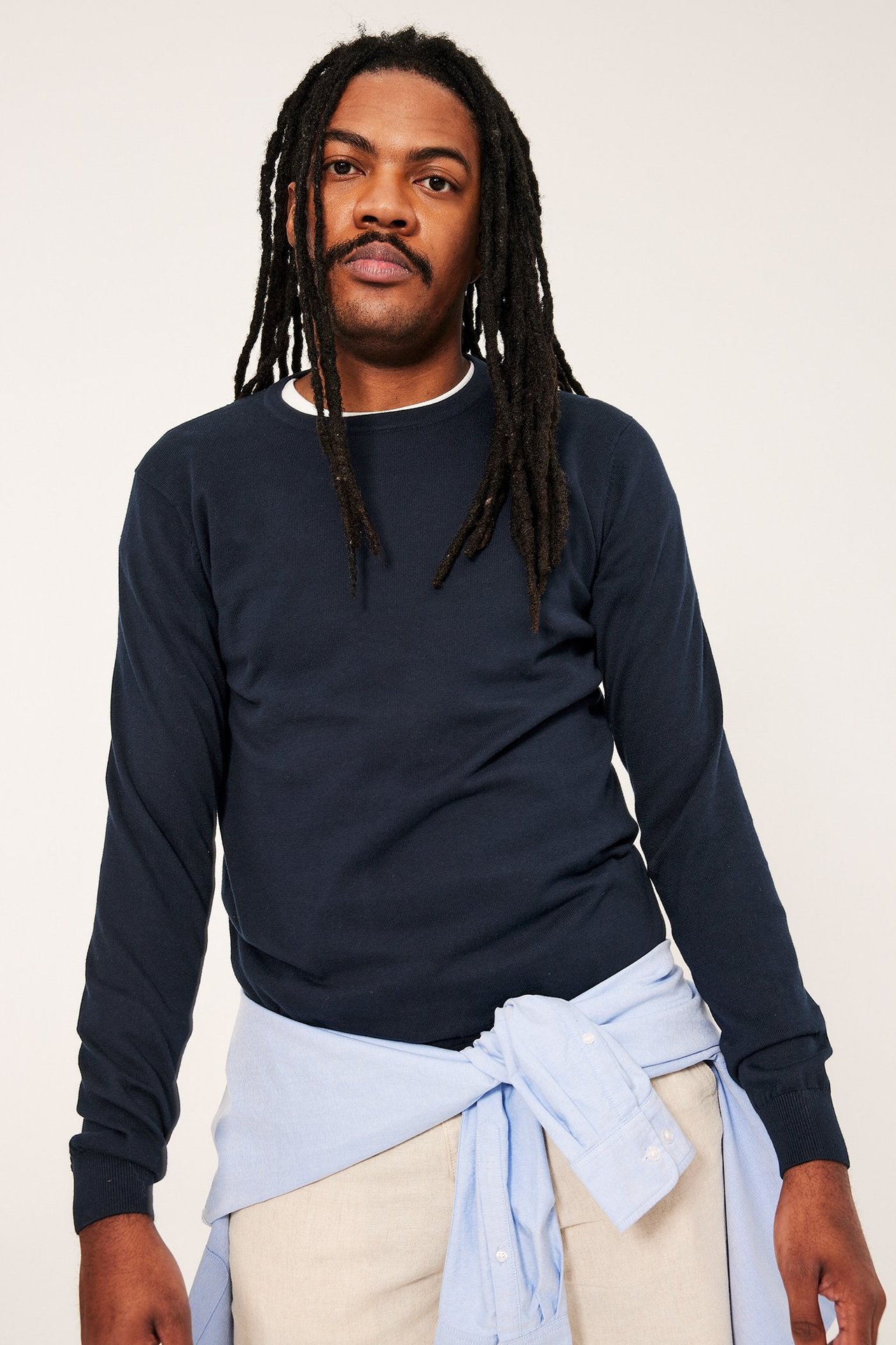 Frontvy: Man i mörkblå finstickad bomullströja, vit t-shirt, ljusblå skjorta (midja), beiga byxor.
