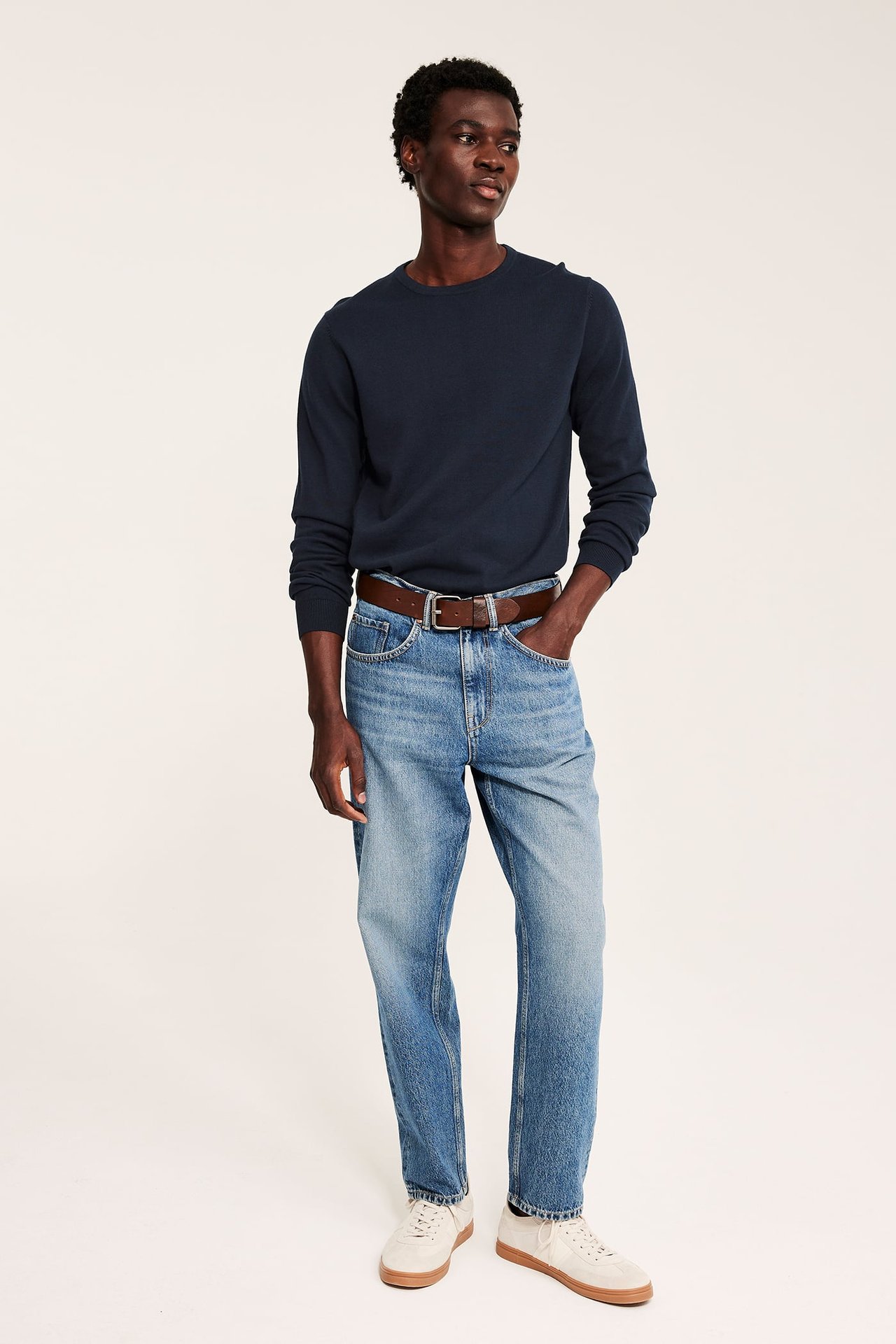 Forfra: Mann i mørkeblå finstrikket bomullsgenser, lyseblå jeans, brunt belte og beige sneakers.