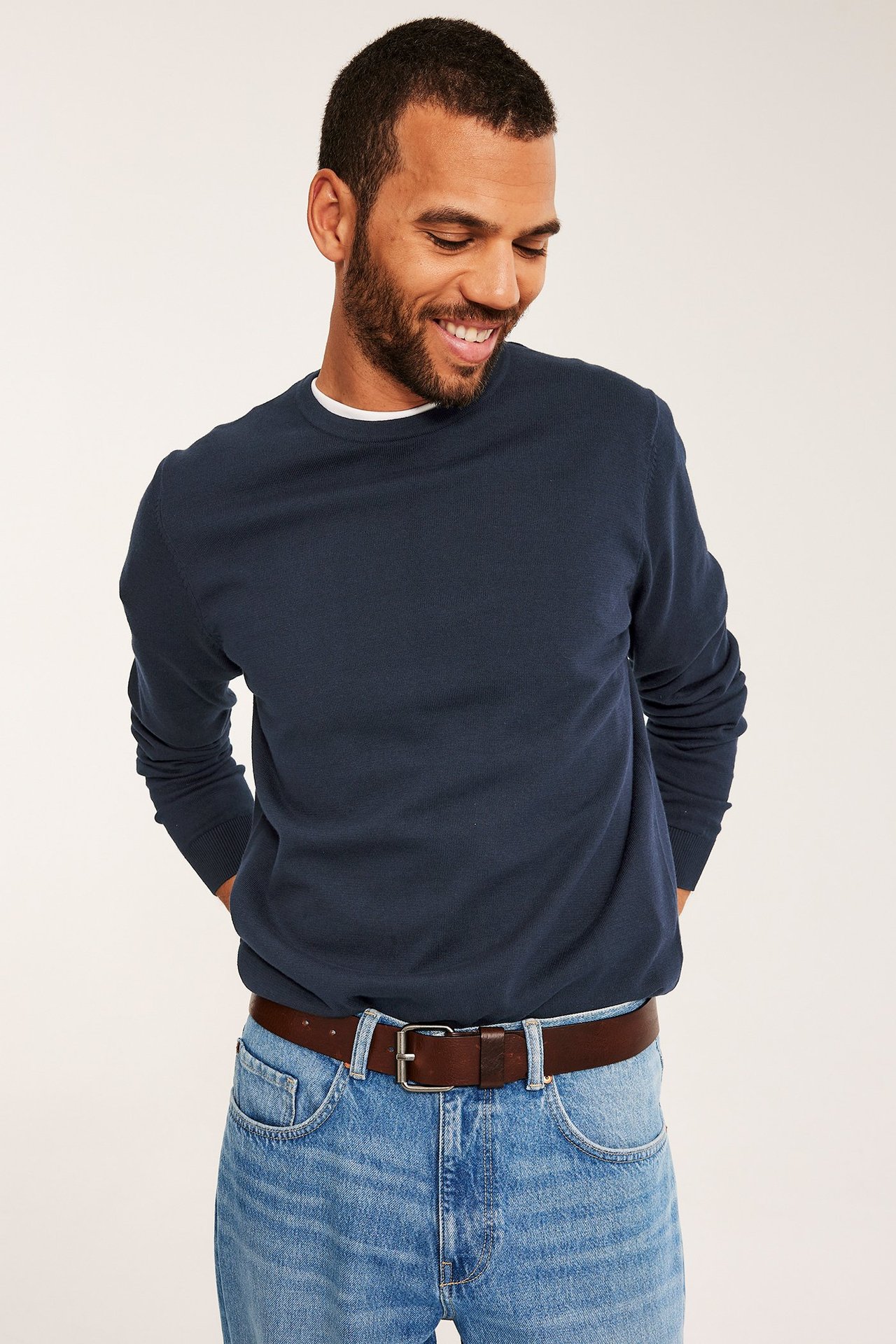 Edestä: mies tummansinisessä hienoneulotussa puuvillaneuleessa, valkoisessa t-paidassa, vaaleansinisissä farkuissa, ruskeassa vyössä.