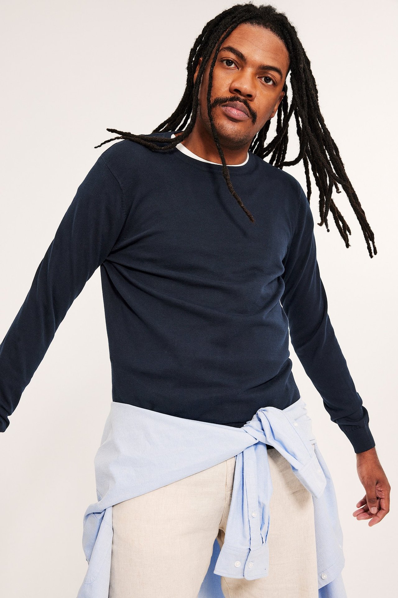 Widok z przodu, mężczyzna w ciemnoniebieskim swetrze, beżowych spodniach, jasnoniebieska koszula w pasie.