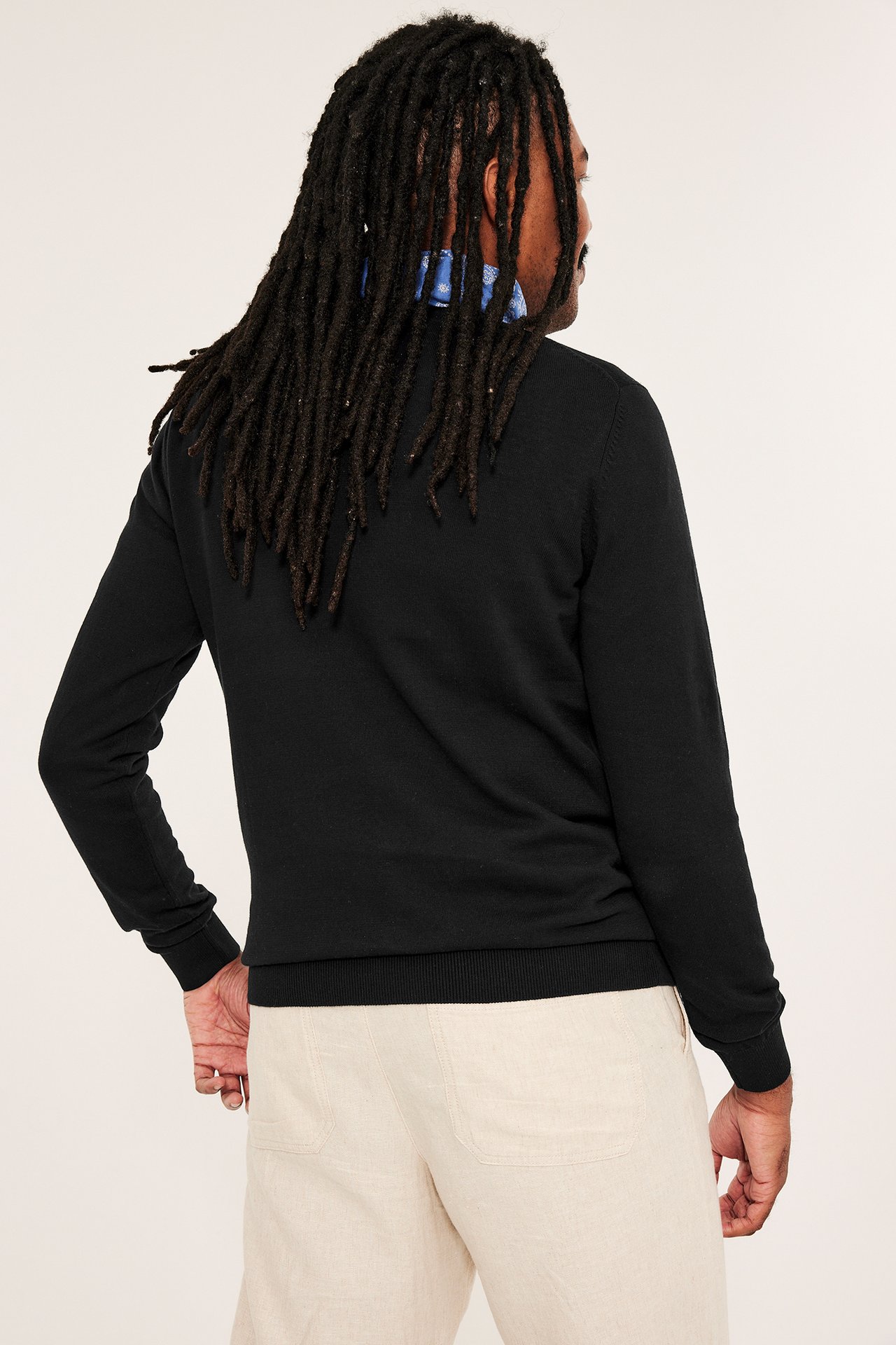 Mężczyzna, widok z tyłu, w czarnym, cienkim bawełnianym swetrze, jasnobeżowych spodniach i niebieskiej bandanie.