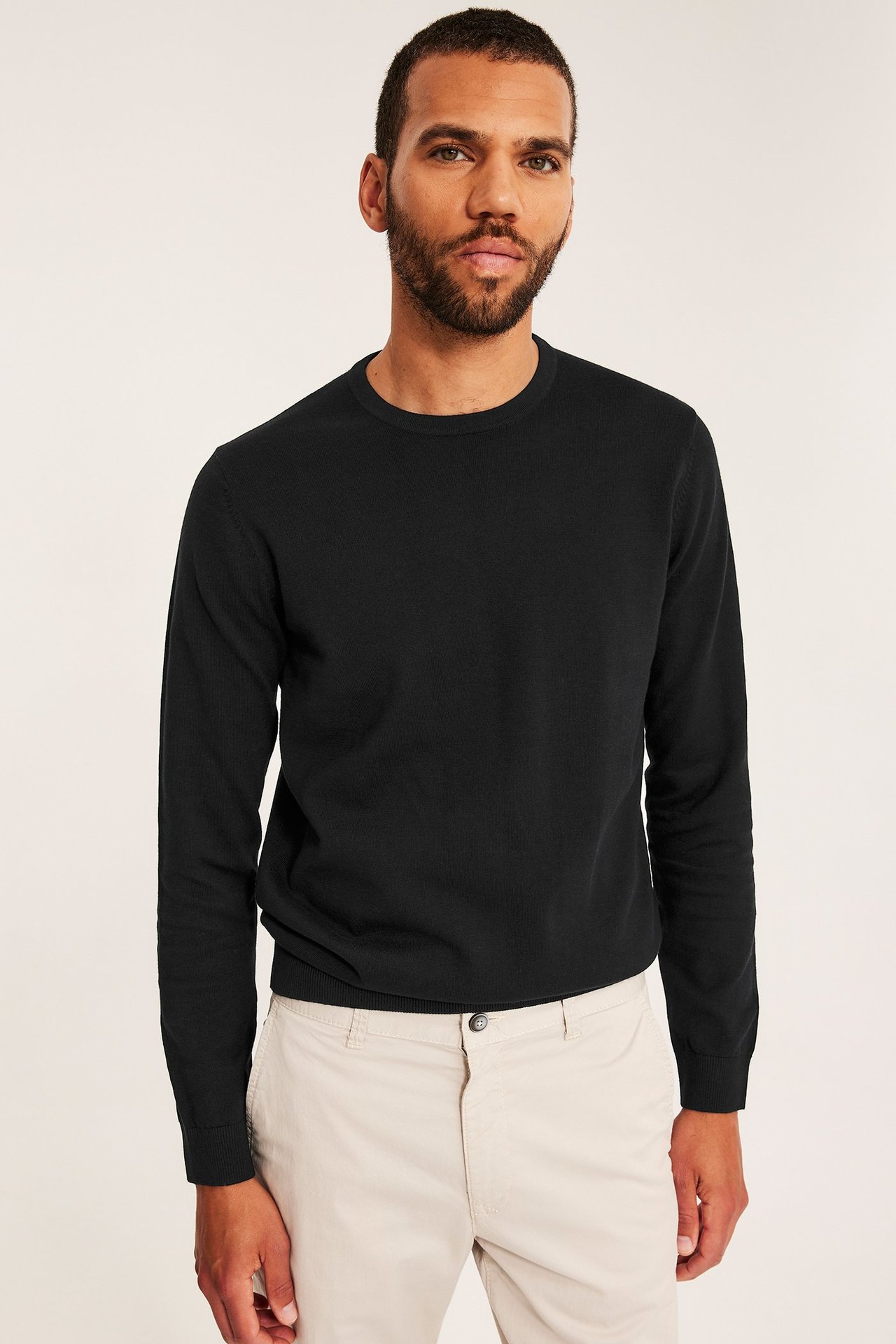 Widok z przodu: Mężczyzna w czarnym, drobno dzianinowym bawełnianym swetrze i jasnobeżowych spodniach.