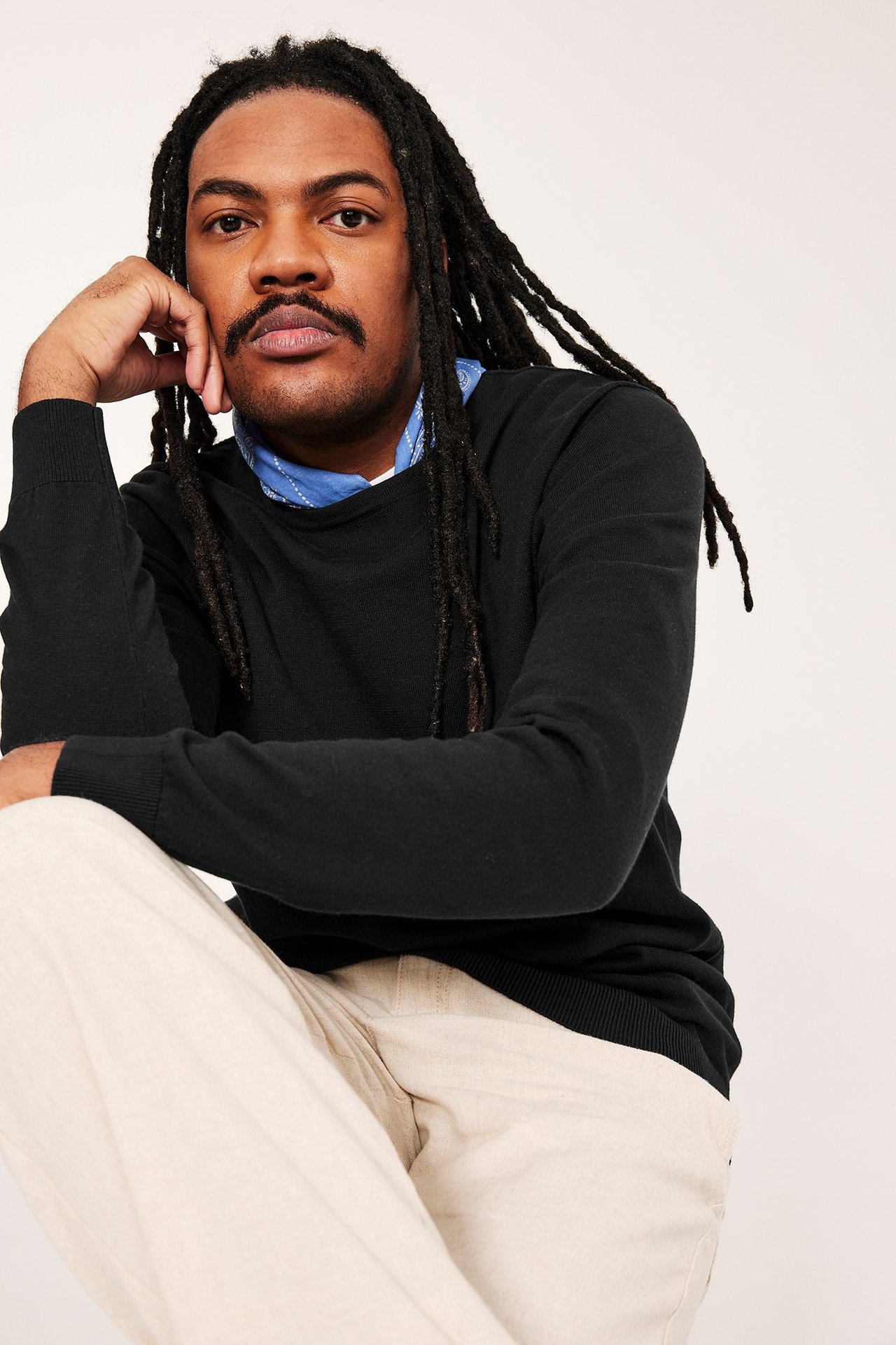 Widok z przodu: mężczyzna w czarnym, dzianym bawełnianym swetrze, białej koszuli, beżowych spodniach i niebieskiej bandanie.