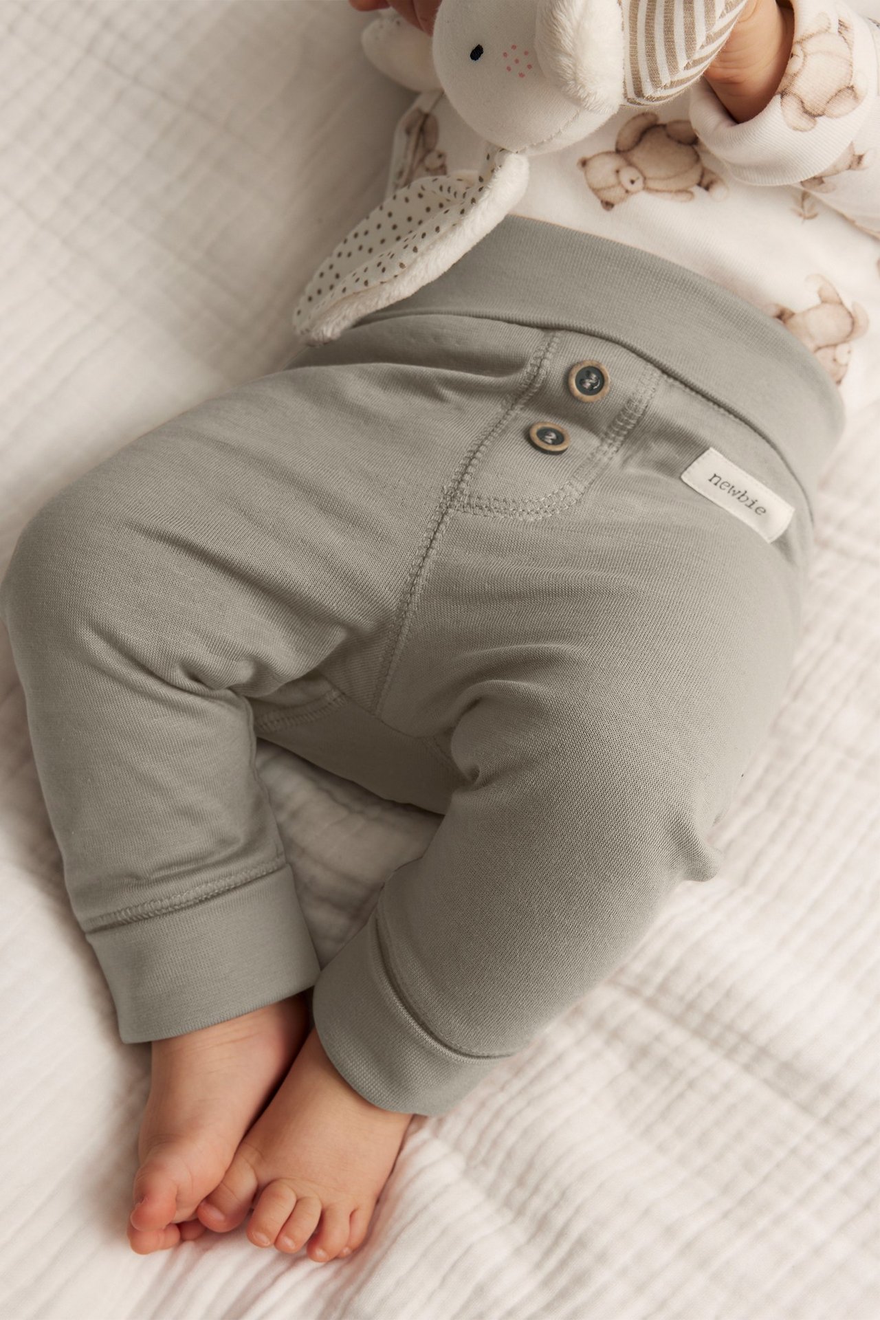 Närbild på baby i beiga bomullsleggings med dekorativa knappar, håller en vit gosedjurskanin.