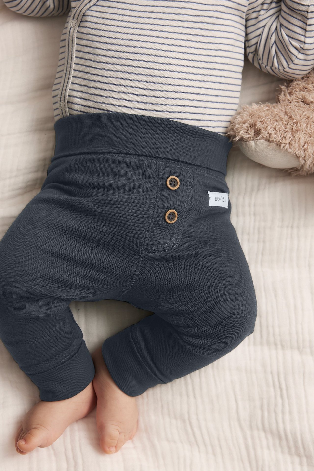 Uppifrån-vy av en baby i mörkblå bomullsleggings med träknappar och en randig body.