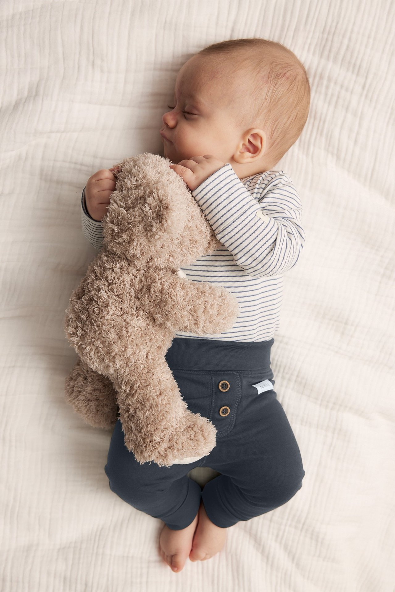 Yläkuva nukkuvasta vauvasta tummansinisissä nappileggingseissä, raidallisessa bodyssä, pitäen ruskeaa nallekarhua.