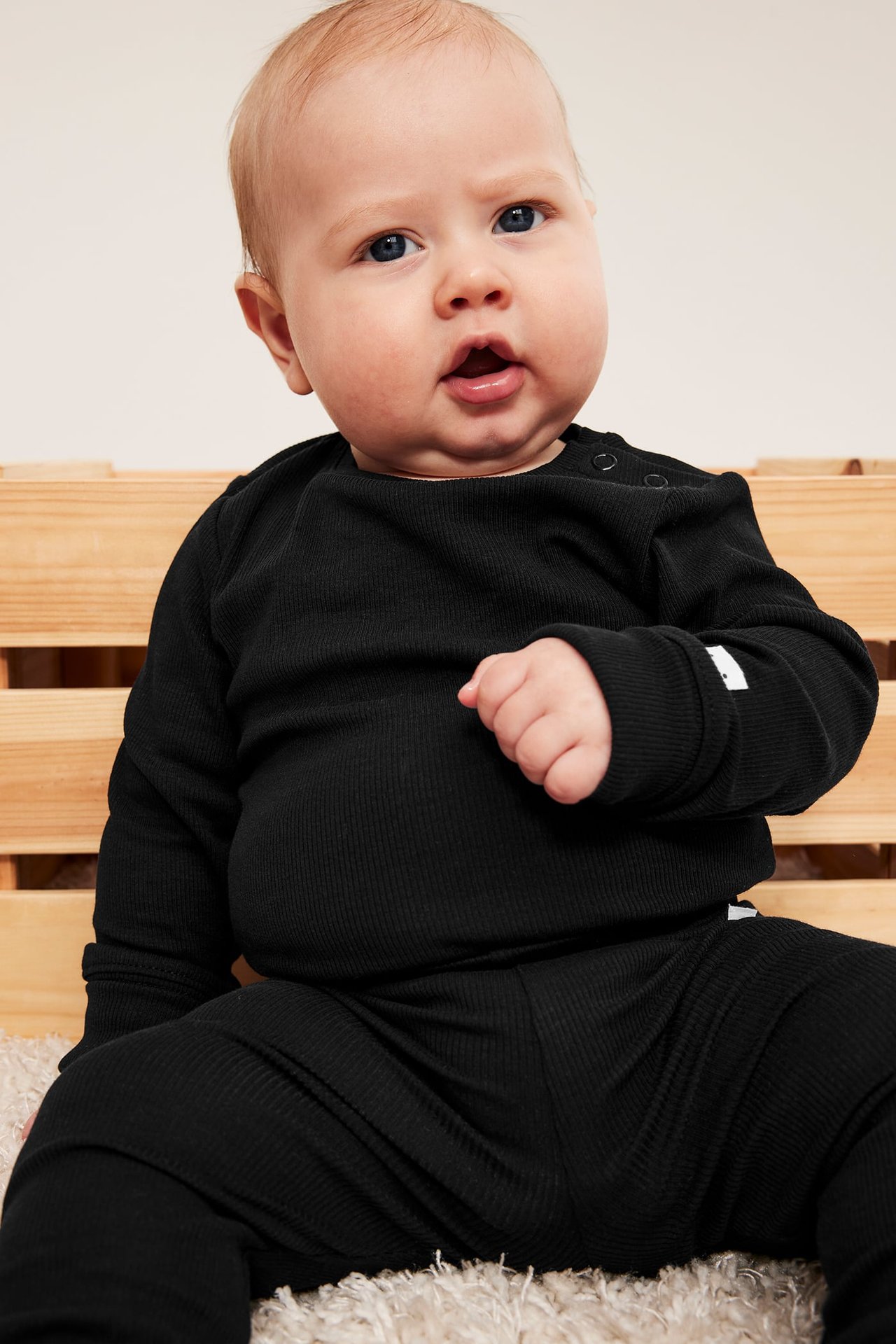 Forfra en baby i svart ribbet langermet topp med skulderknapper og matchende ribbet bukse.