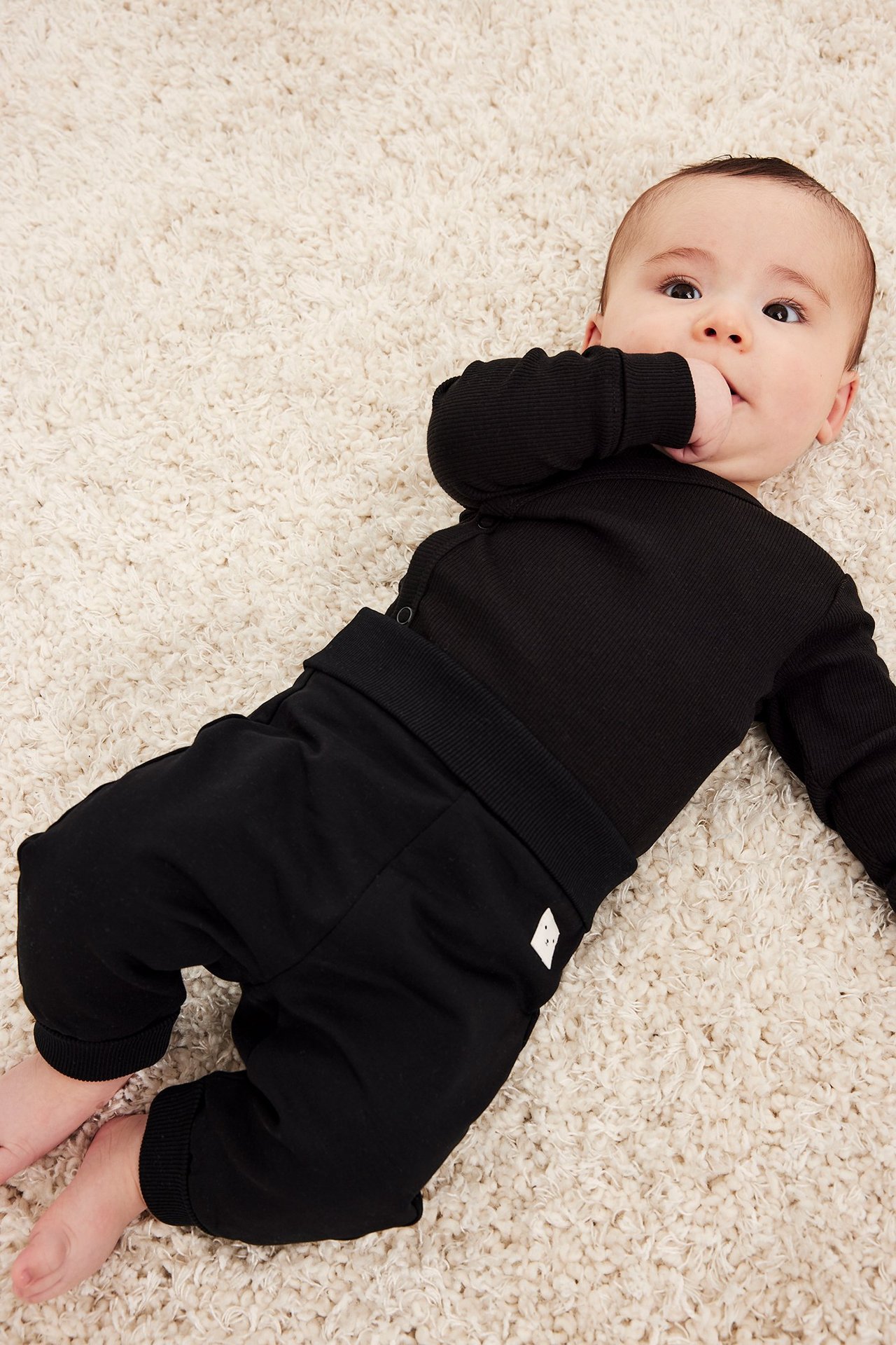 Ylhäältä näkymä vauvasta mustassa ribatussa pitkähihaisessa kietaisubodyssa ja mustissa housuissa.