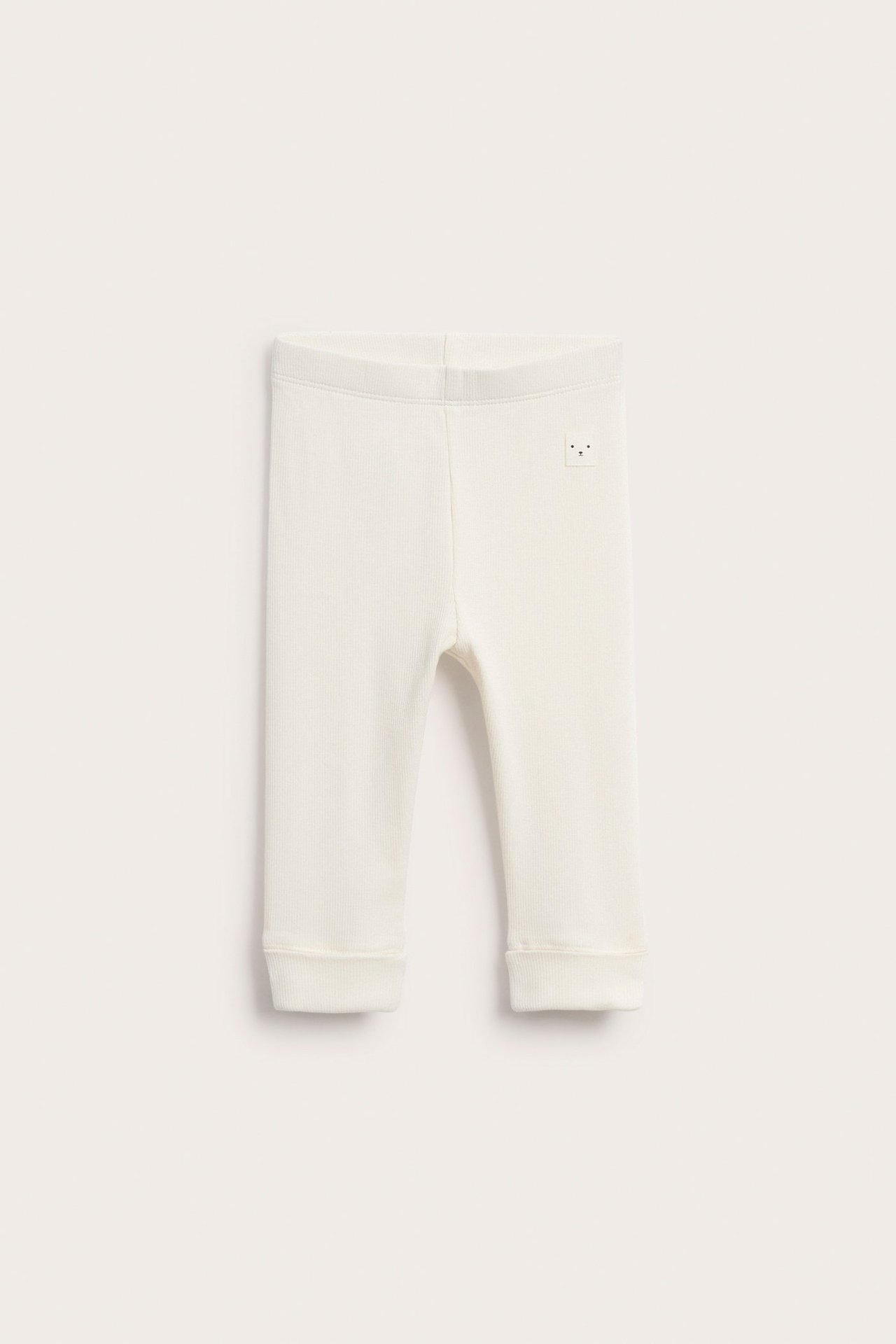 Widok z przodu kremowych, prążkowanych legginsów niemowlęcych z szerokim pasem, mankietami i małą naszywką z buźką.
