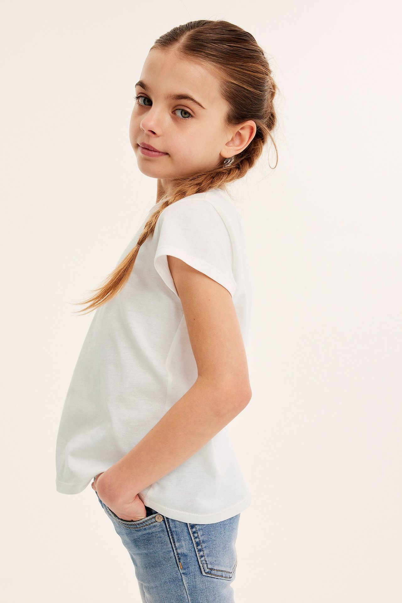 Widok z boku dziecka w kremowym bawełnianym T-shircie i niebieskich dżinsach z rękami w kieszeniach.
