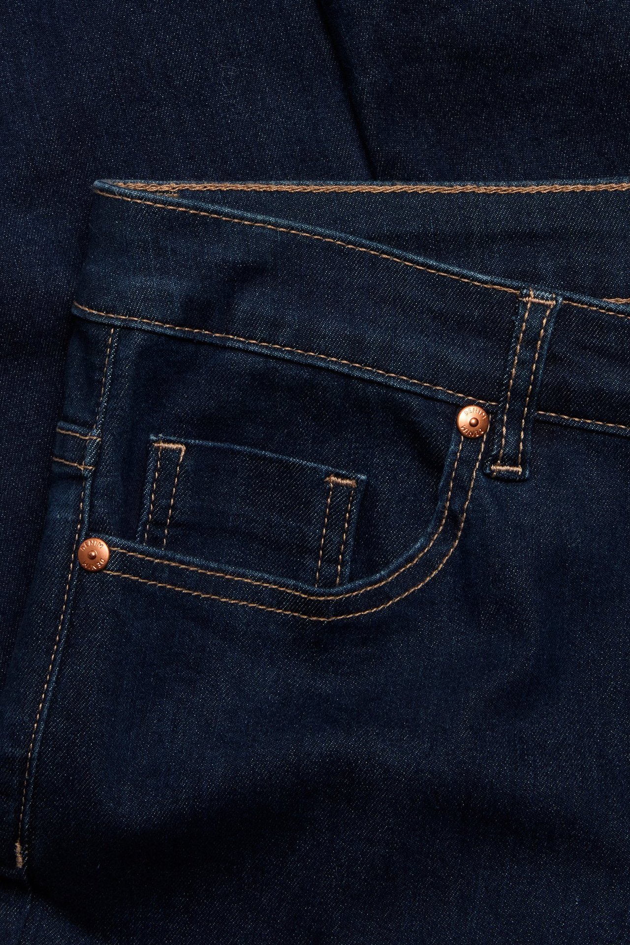 Zbliżenie na ciemne jeansy straight z jasnobrązowymi szwami i miedzianymi nitami na przedniej kieszeni.