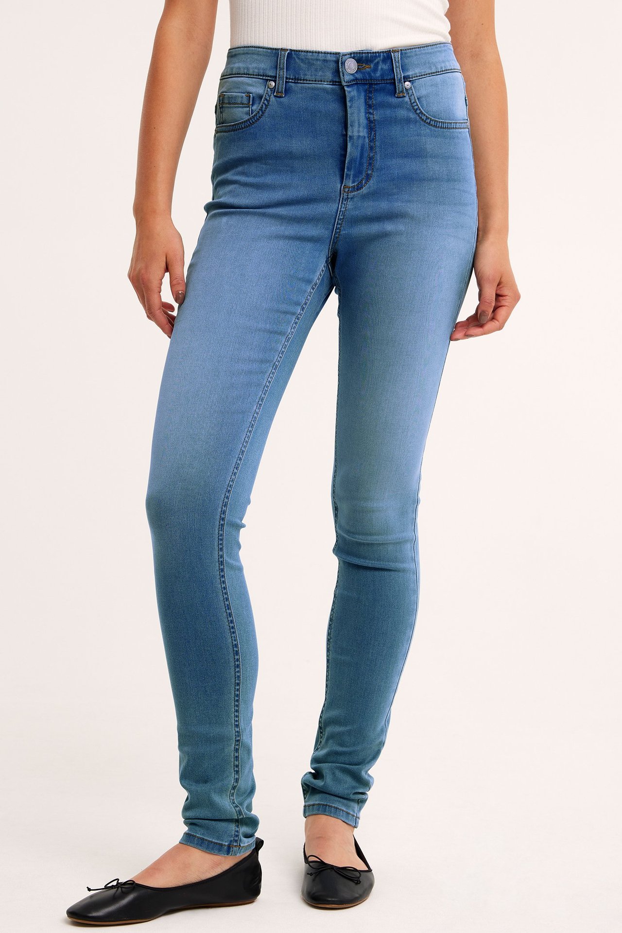 Framifrån, kvinna i ljusblå supersmala denimjeans, vit ribbad topp och svarta ballerinaskor.