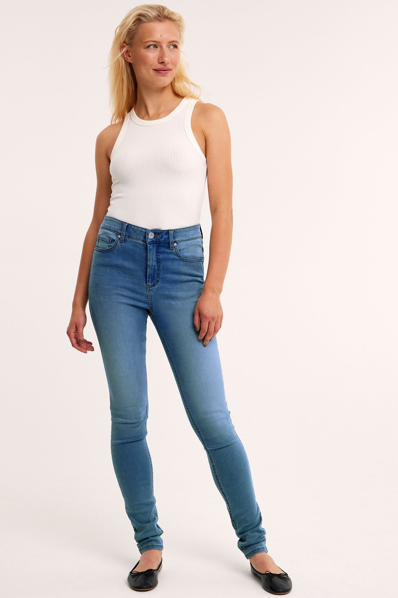 Framifrån, kvinna i ljusblå supersmala denimjeans, vit ribbad tanktopp och svarta ballerinaskor.