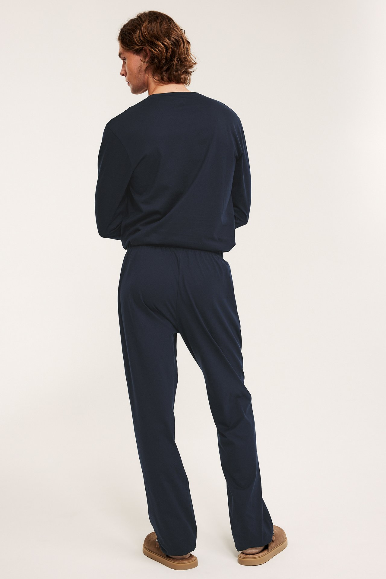 Mężczyzna z tyłu: granatowe piżamowe spodnie dresowe, pasująca koszulka i brązowe kapcie.