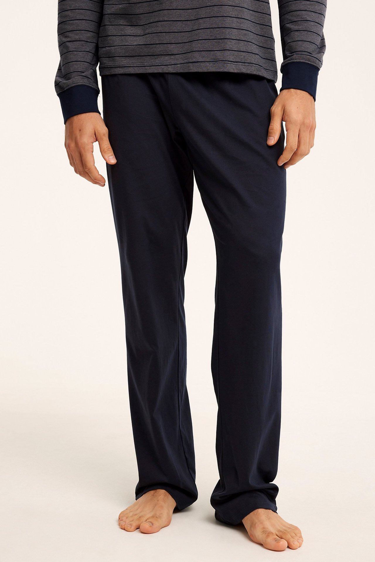 Edestäpäin: tummansiniset puuvillajersey-pyjamahousut, joita mies käyttää harmaaraidallisen paidan kanssa.